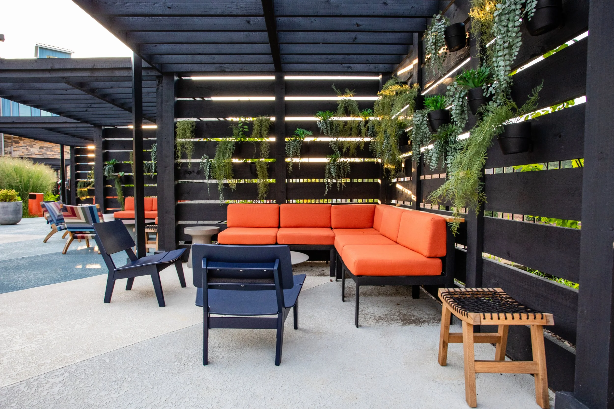 Outdoor seating area with orange cushioned sectional sofa, black chairs, small tables, and a wooden stool, enclosed by a black wooden slat privacy wall decorated with hanging greenery.