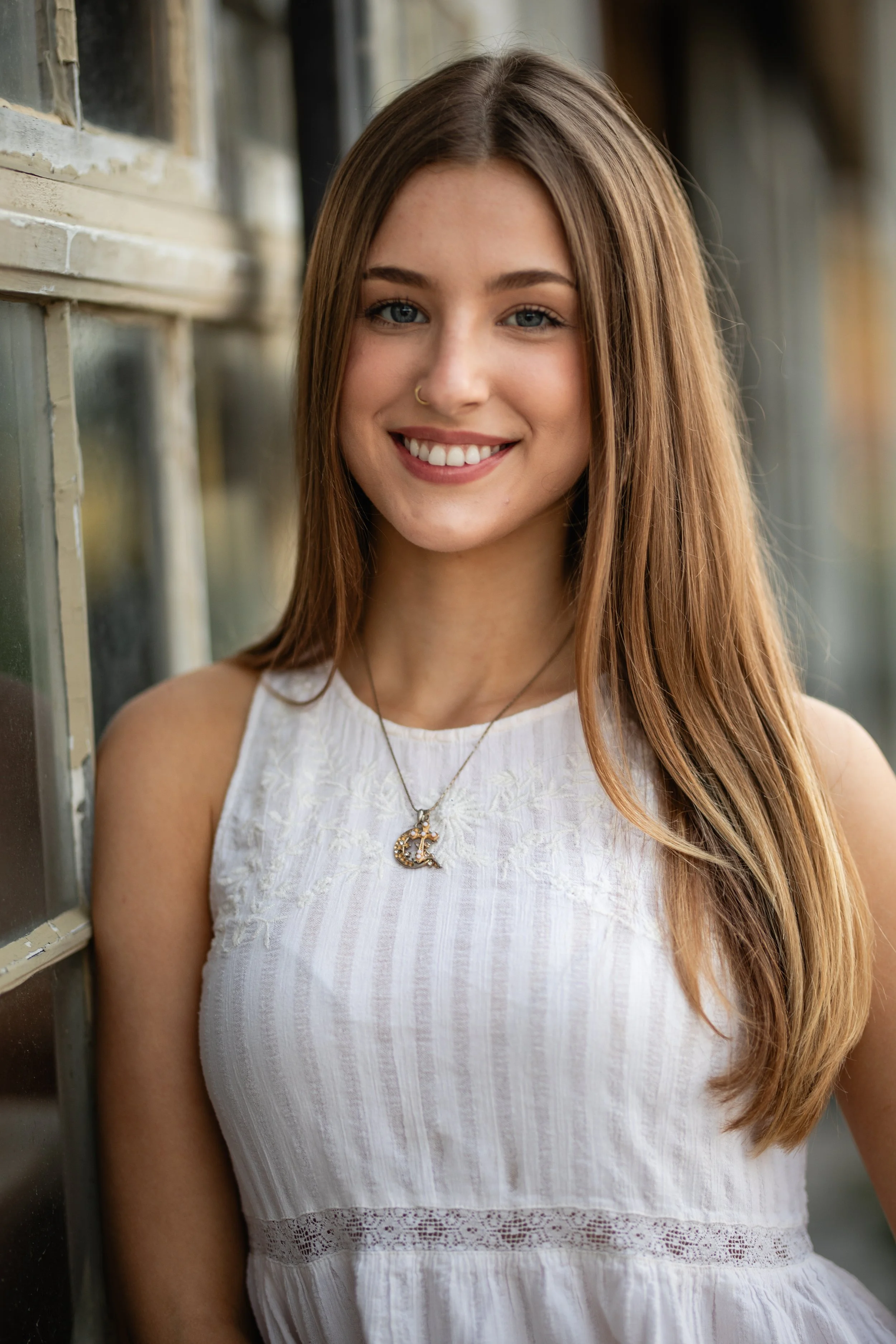 A young woman with long, light brown hair and blue eyes, smiling, wearing a white sleeveless top and a necklace with a moon-shaped pendant, standing next to a window.