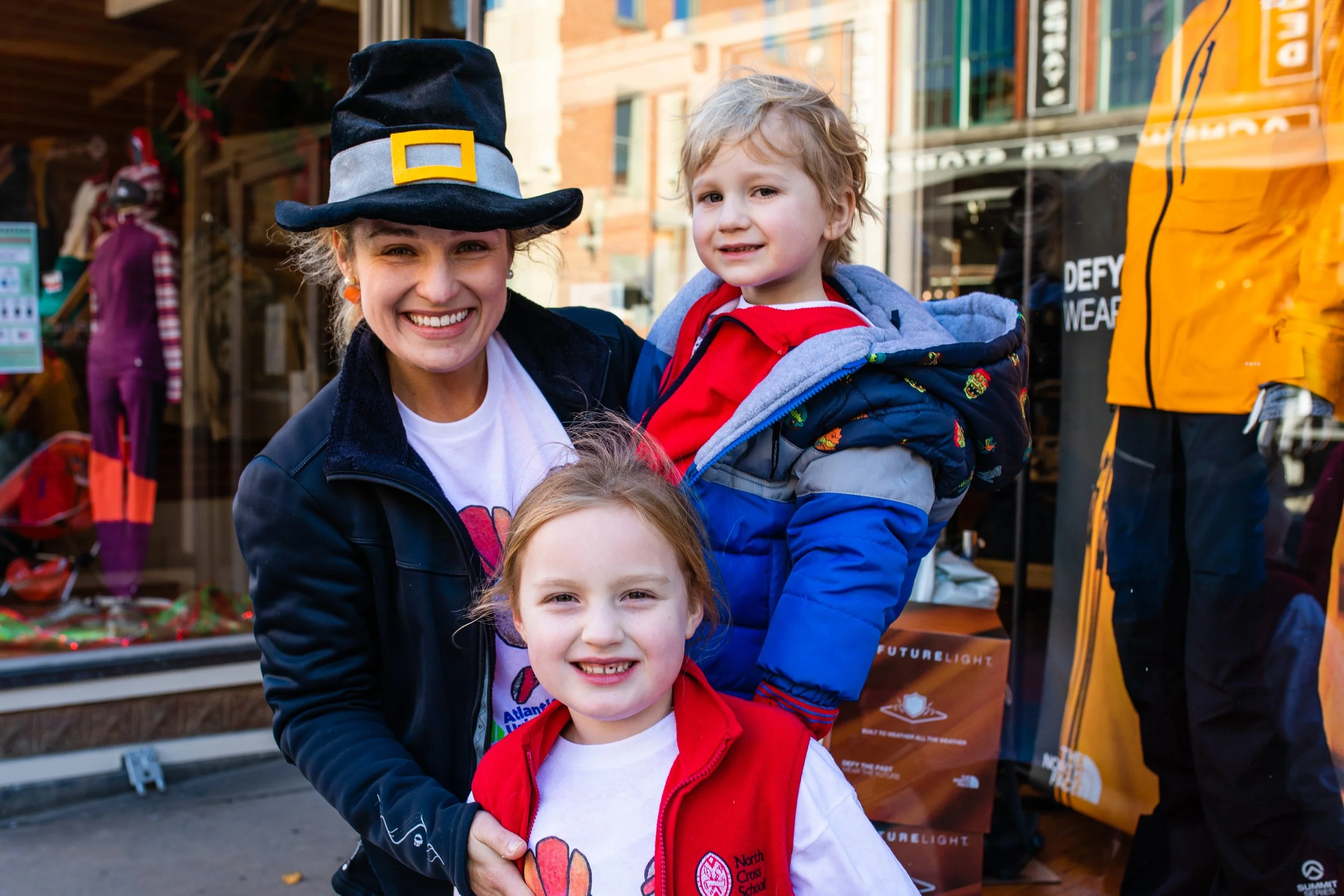 A woman wearing a black hat resembling a pilgrim's hat, smiling and holding two children, one girl with red hair and another boy with blonde hair, in an outdoor setting with store windows and mannequins in the background.