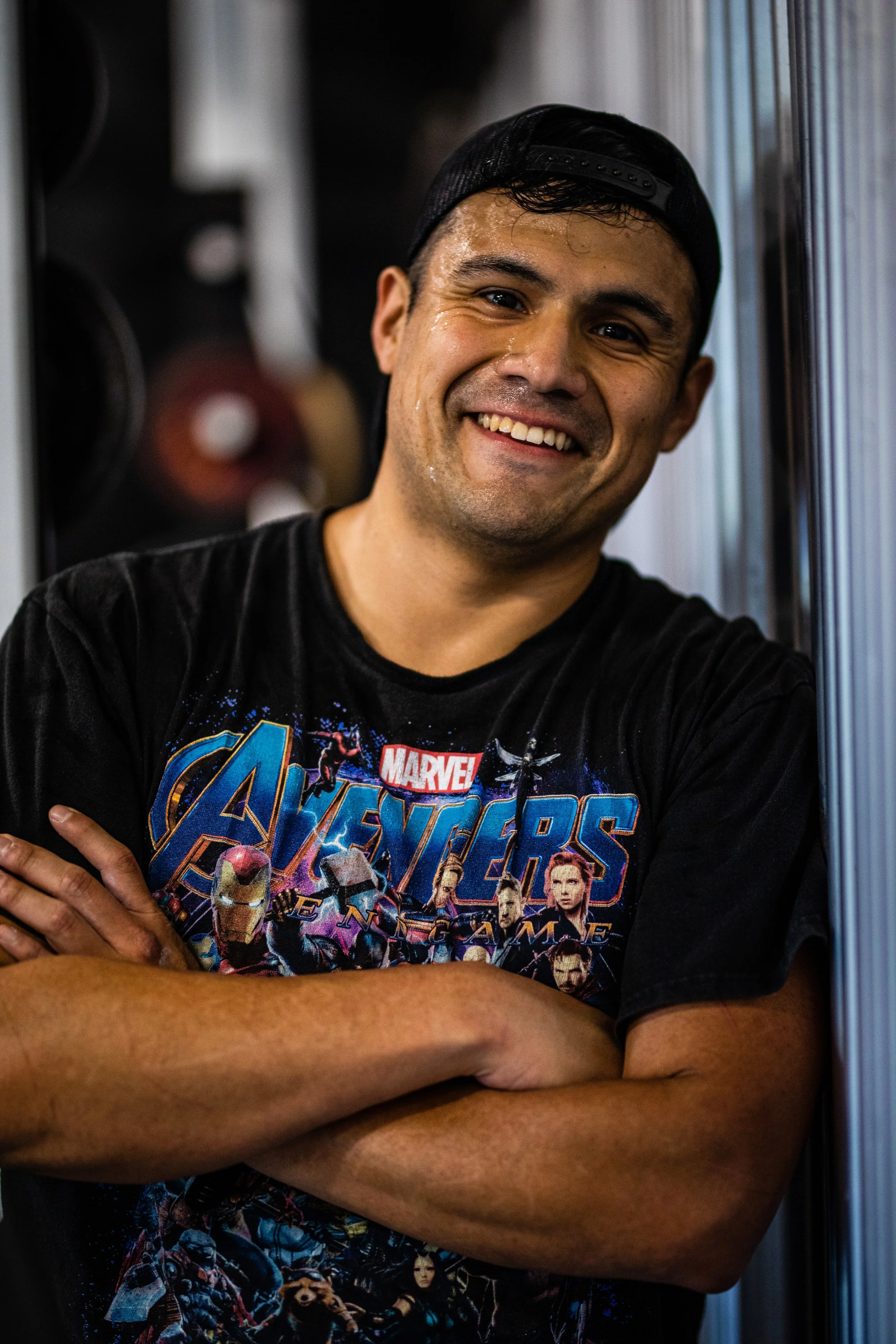 A young man wearing a Marvel Avengers t-shirt and a black cap, smiling with his arms crossed, standing indoors near a metal wall.