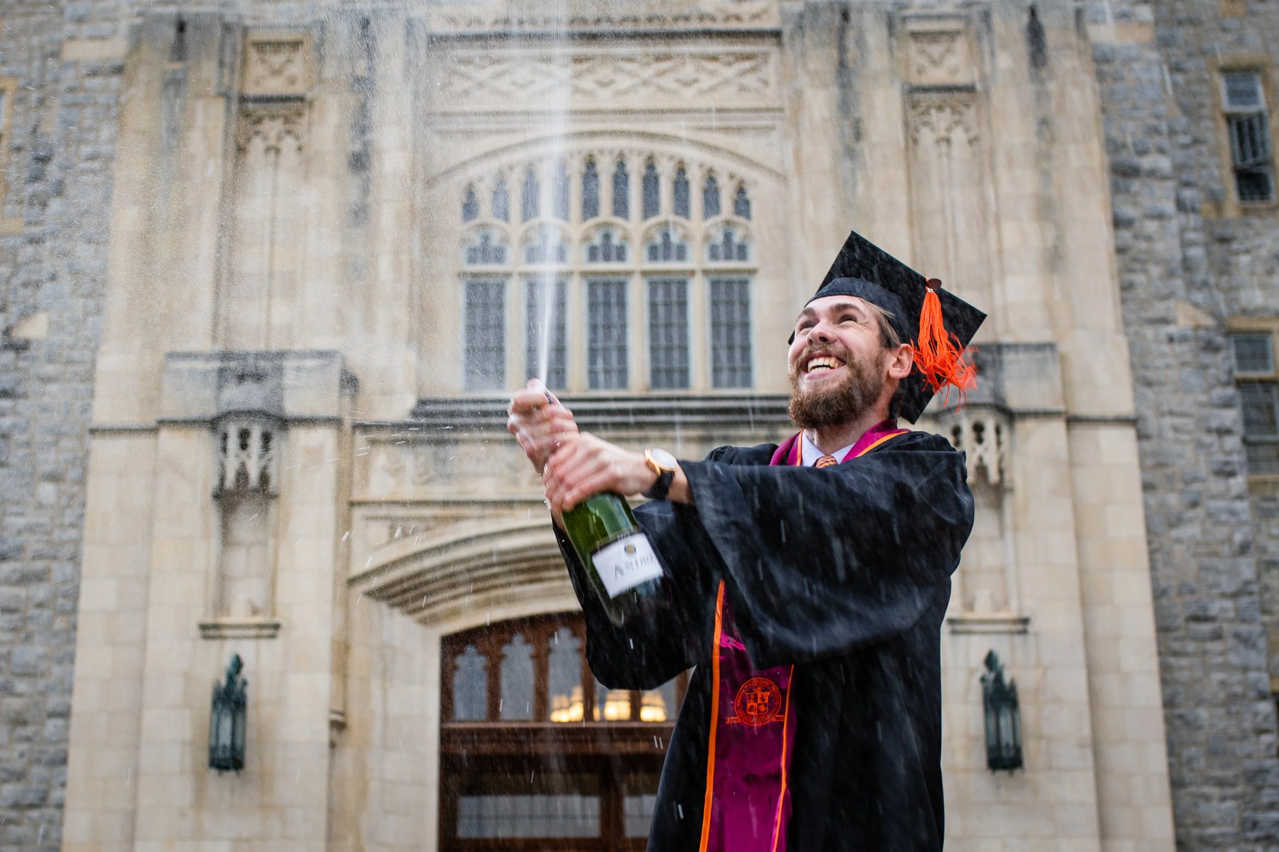 A man celebrating in graduation attire popping a champagne bottle outside a stone building.