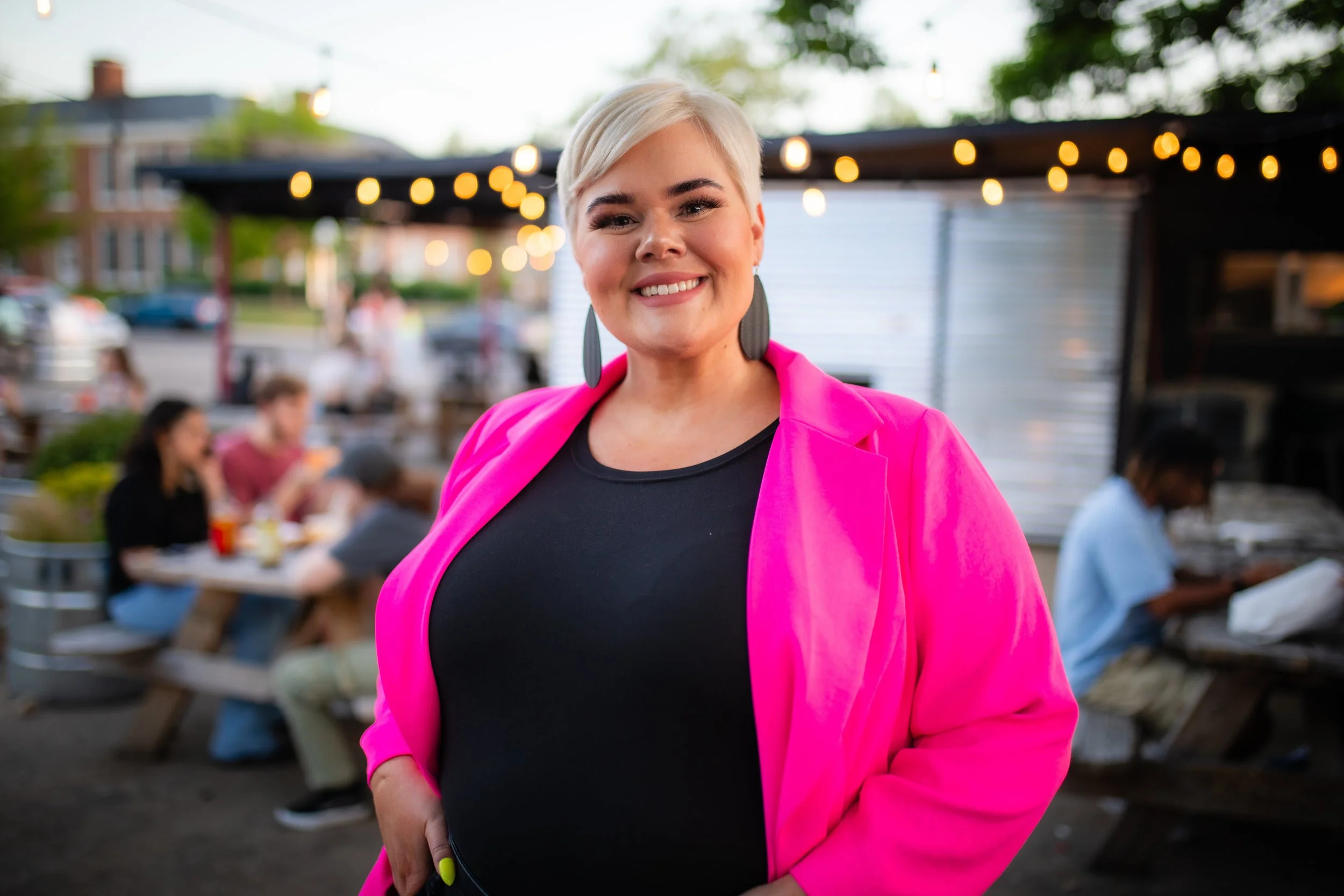 A woman with short blonde hair smiling at the camera, wearing a hot pink jacket and black top, stands outdoors at evening with string lights hanging overhead and people sitting at picnic tables in the background.