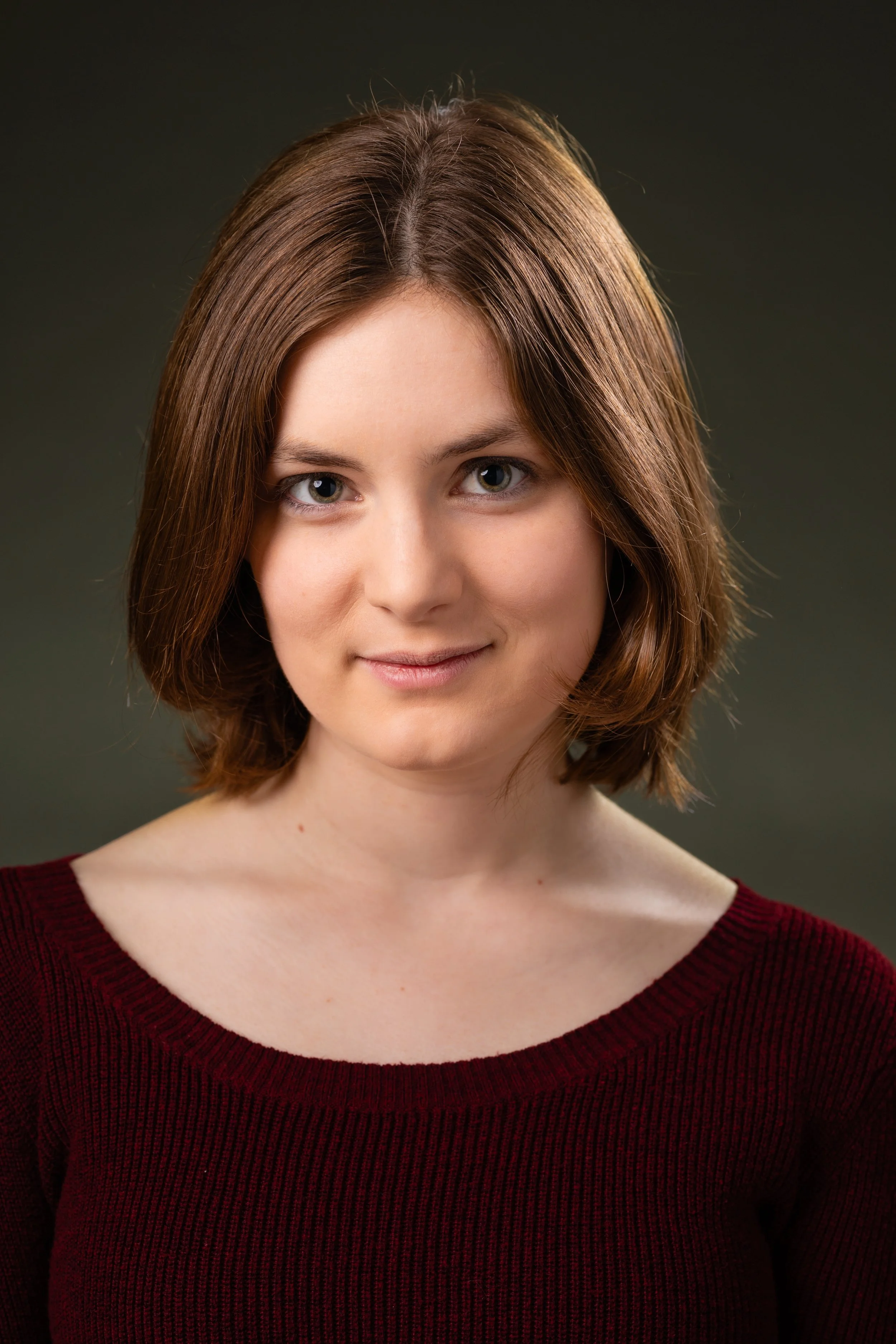 Close-up portrait of a young woman with shoulder-length brown hair, light skin, and blue eyes, smiling softly, wearing a burgundy top, against a dark gradient background.