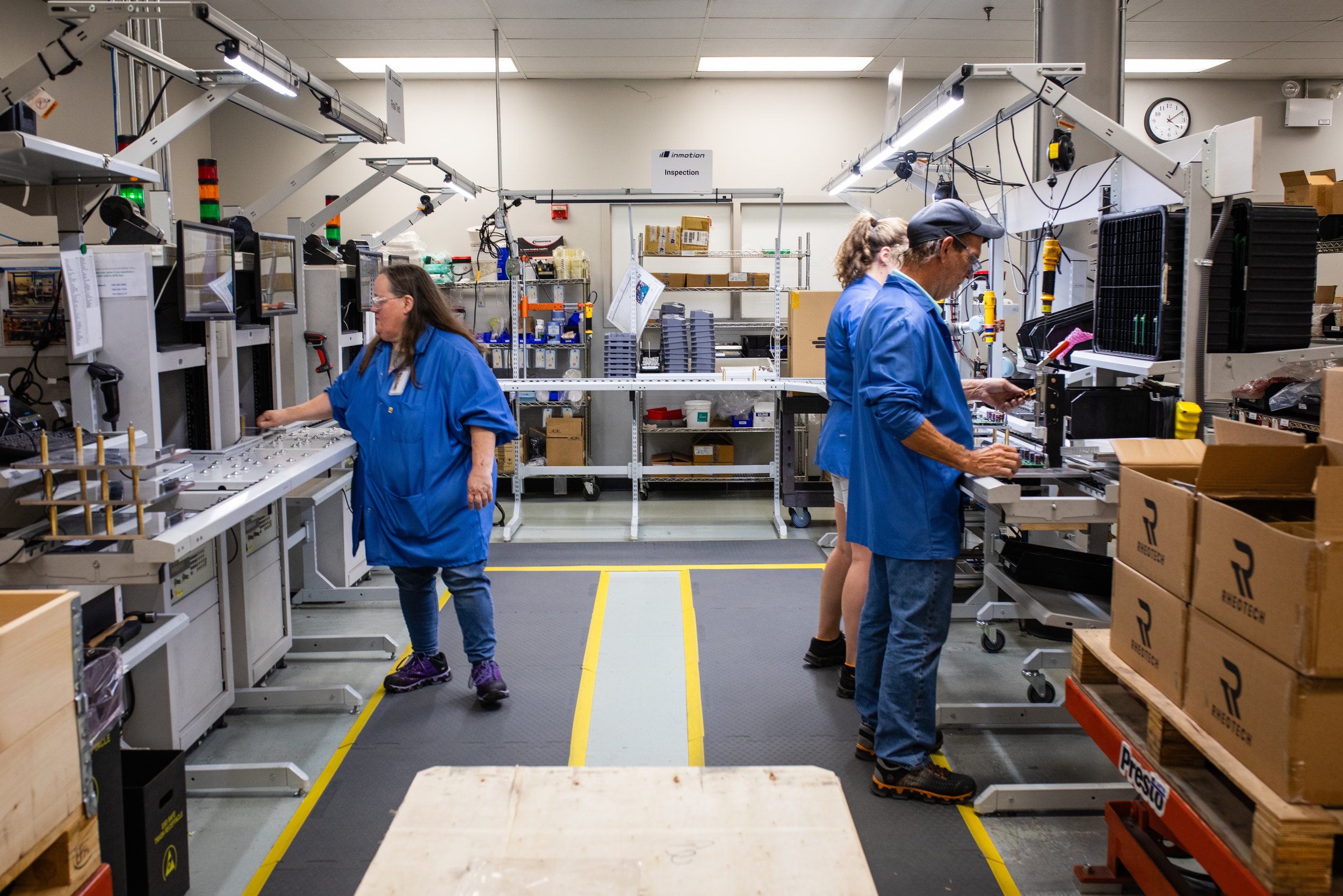 Workers in a manufacturing or assembly factory working at workstations with tools, equipment, and boxes, wearing blue uniforms.