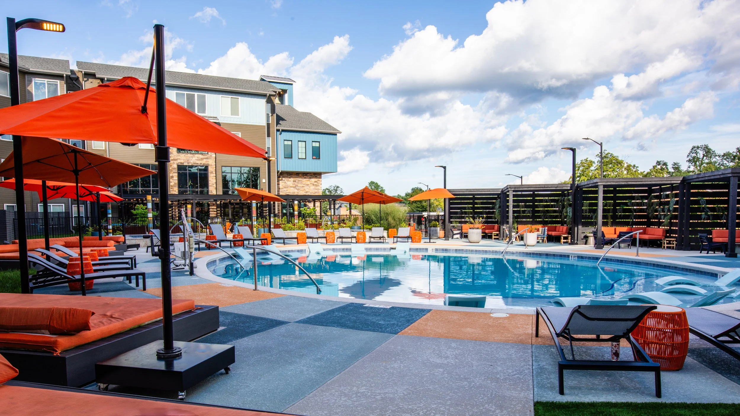 Quiet outdoor swimming pool area with orange umbrellas, lounge chairs, and modern apartment buildings in the background under a partly cloudy sky.