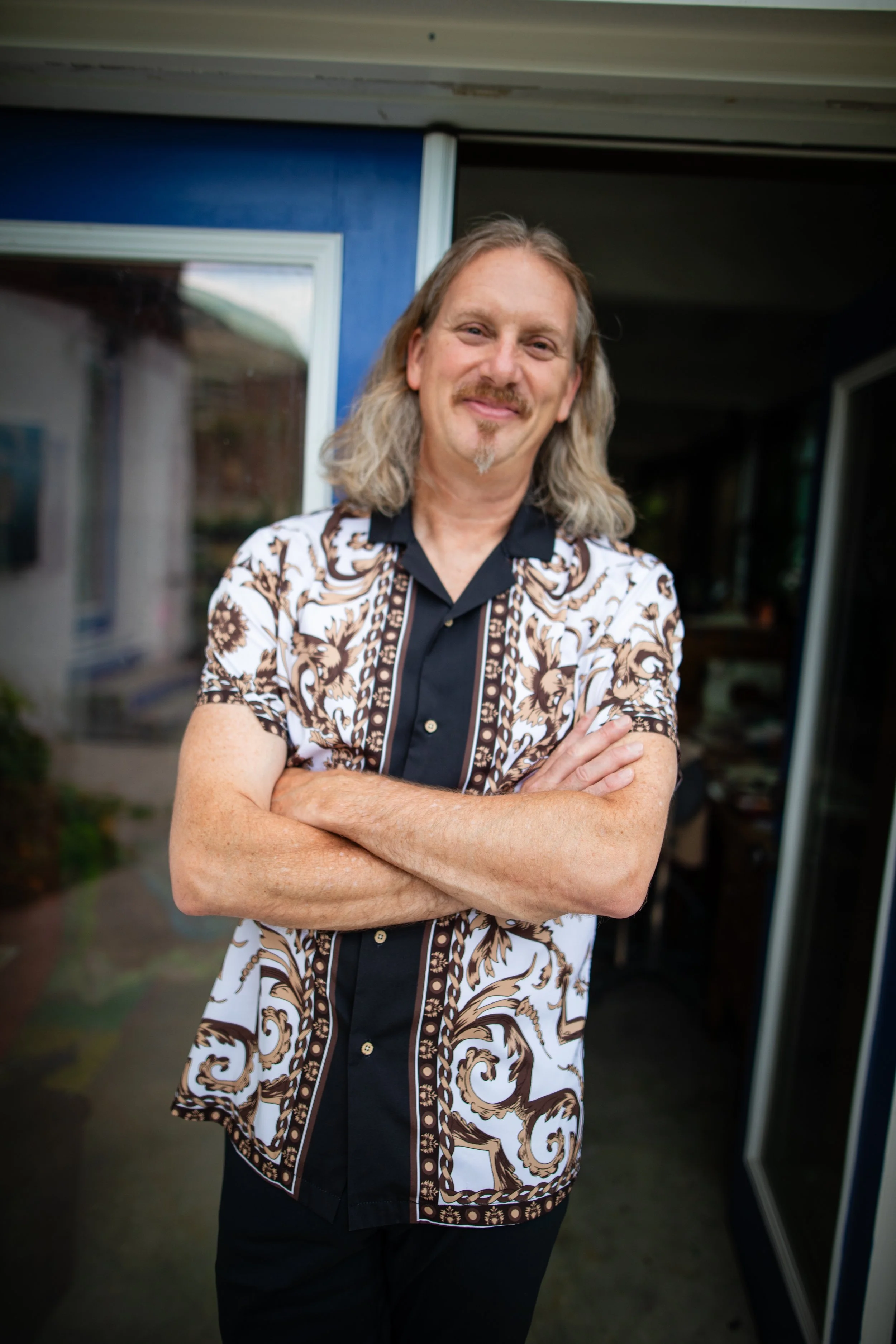 A man with shoulder-length wavy gray hair, a mustache, and goatee smiling with arms crossed, standing outdoors near a blue building with large windows.