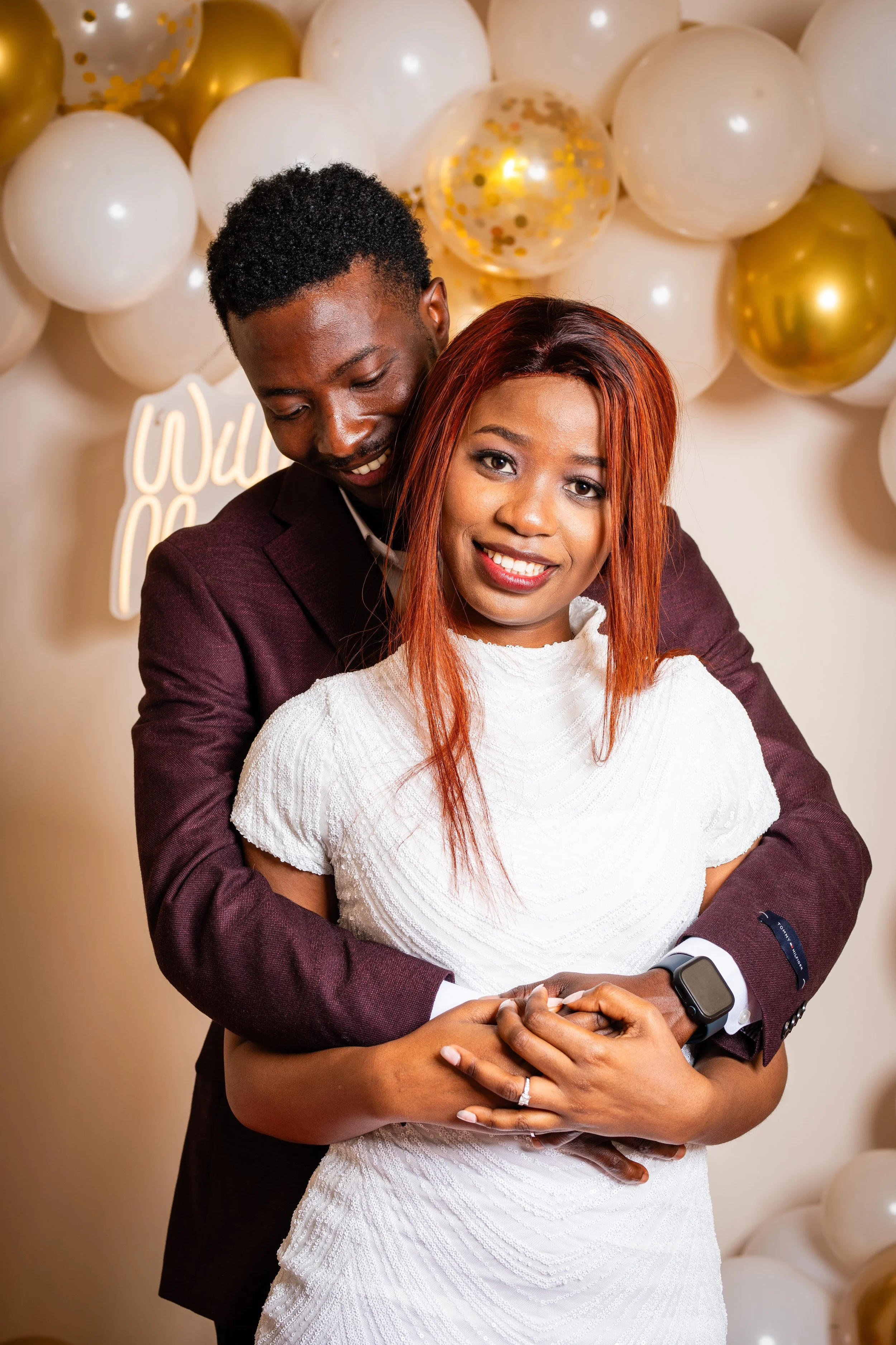 A happy couple hugging at a celebration with white, gold, and clear balloons in the background