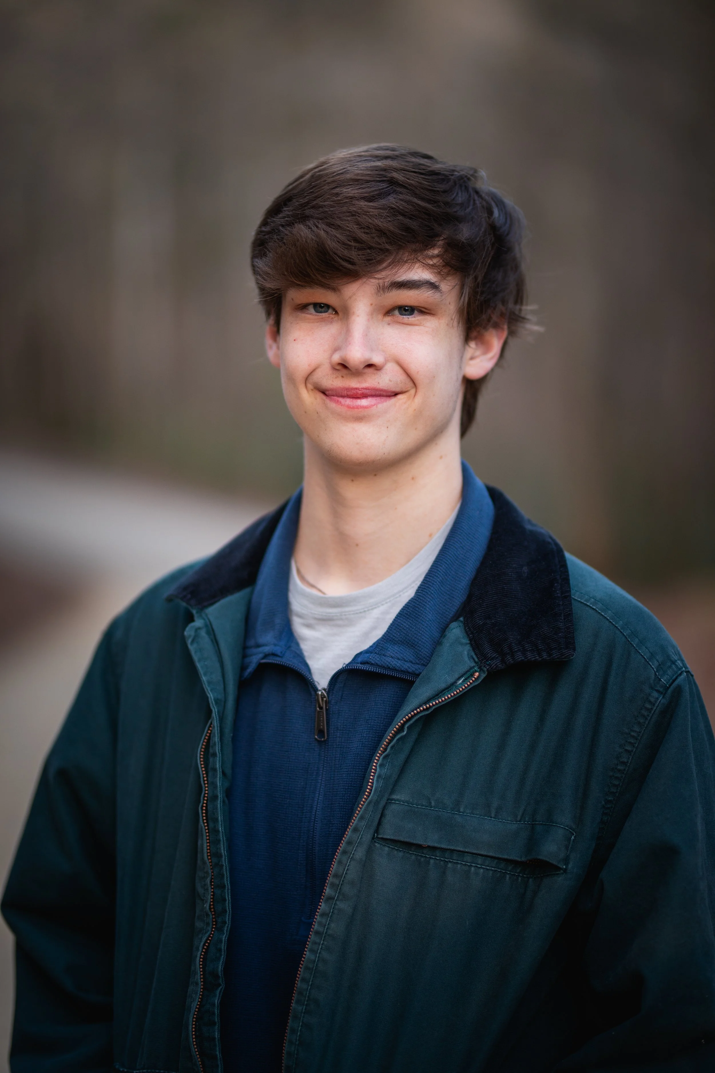 A young man outdoors in a casual jacket, smiling at the camera with a blurred natural background.