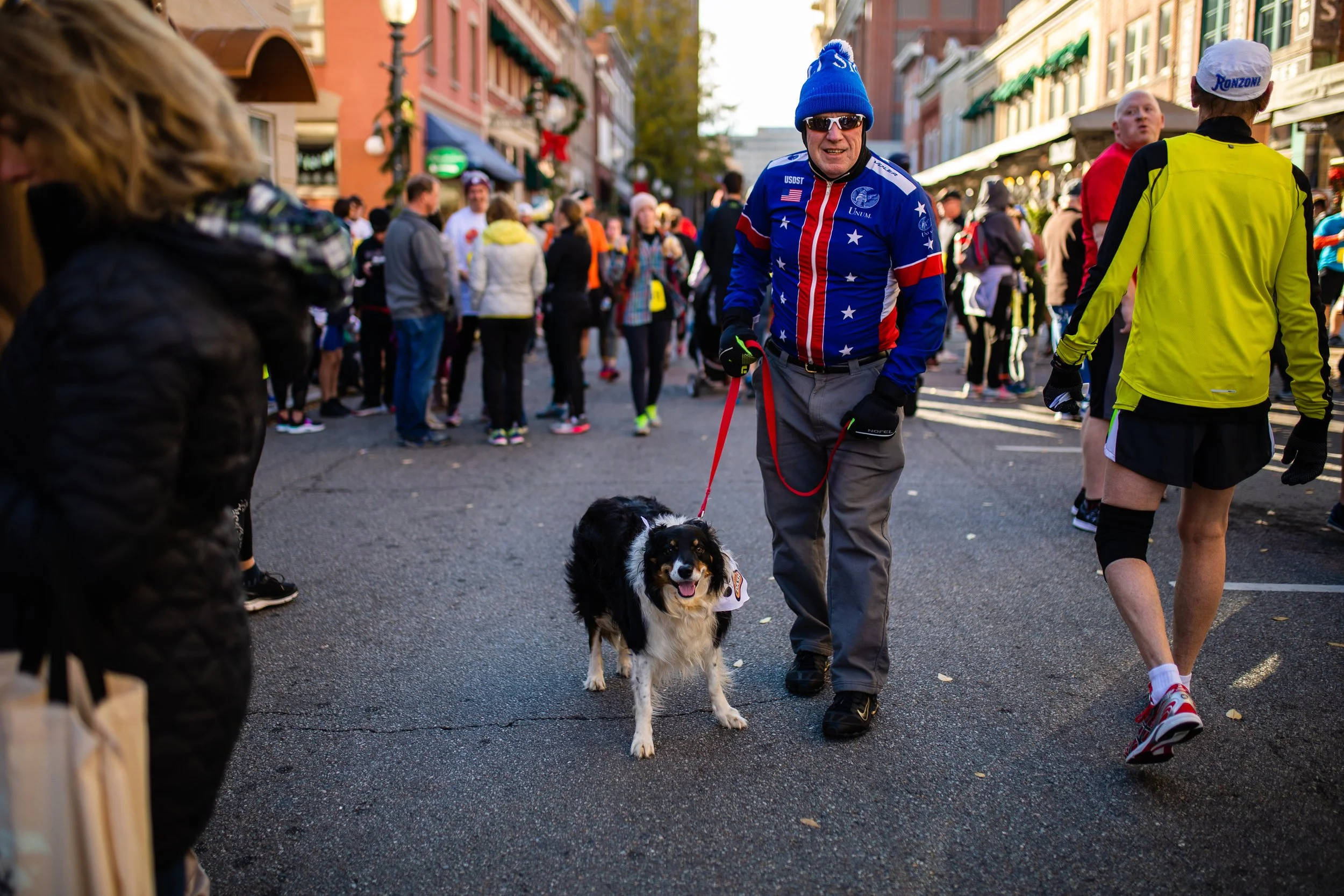 A man walking a dog on a busy city street during a public event or marathon, with several people gathered around and colorful storefronts in the background.