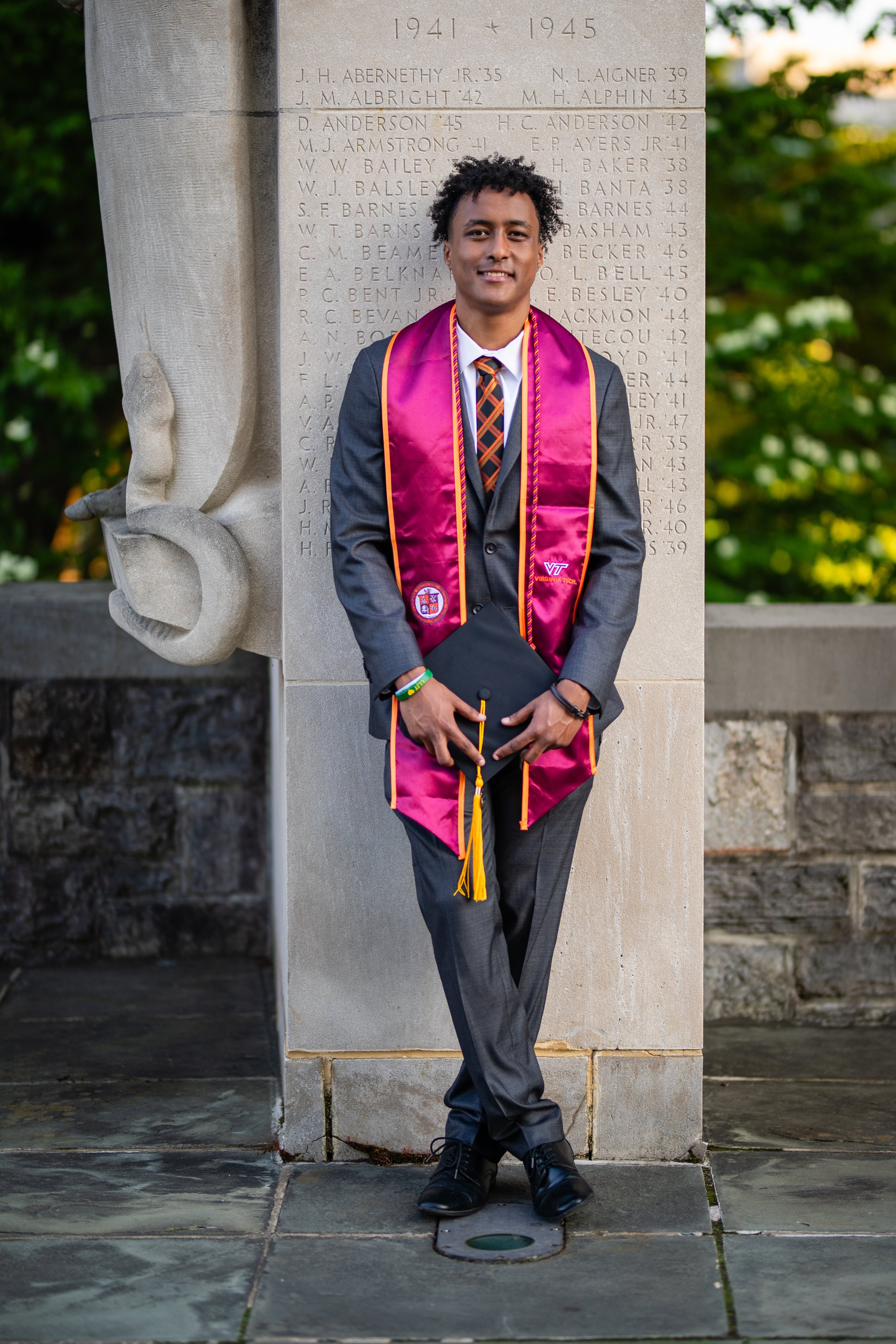 Young man in a gray suit and colorful tie, holding a graduation cap, standing in front of a memorial wall with carved figures, wearing a pink and gold graduation stole, smiling at the camera.