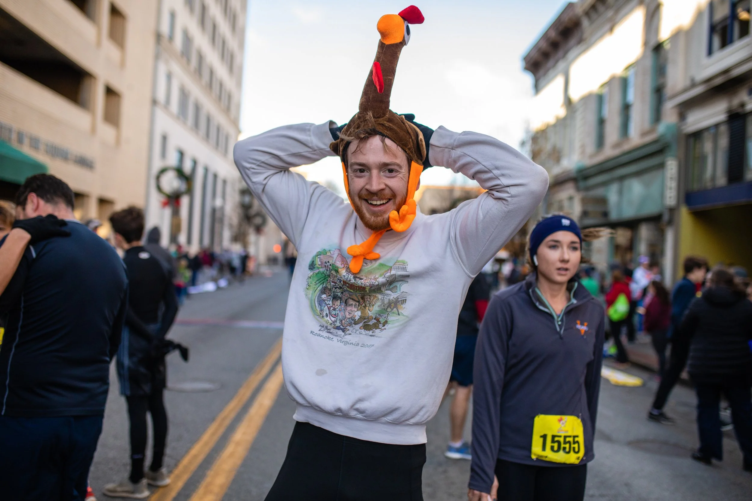 A man wearing a turkey hat, smiling, and adjusting the hat with his hands, participating in an outdoor event with many people around on a city street.