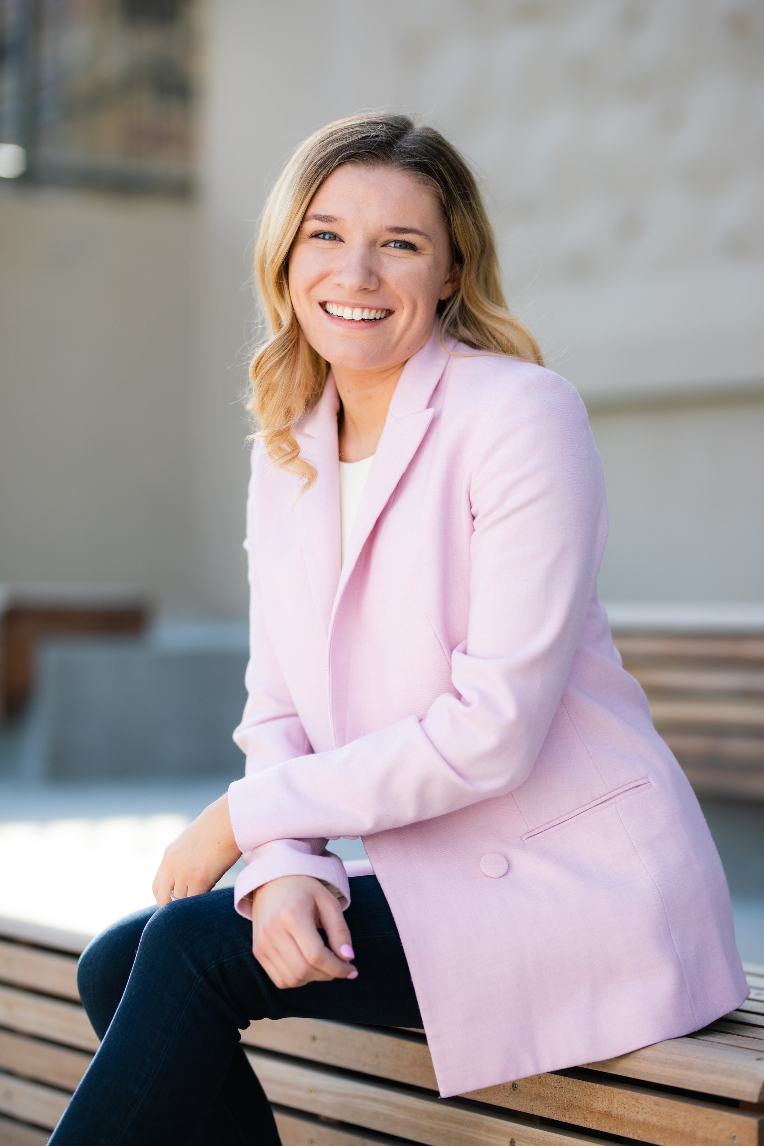 A woman with blonde hair smiling, wearing a light pink blazer and dark jeans, sitting on a wooden bench outside.