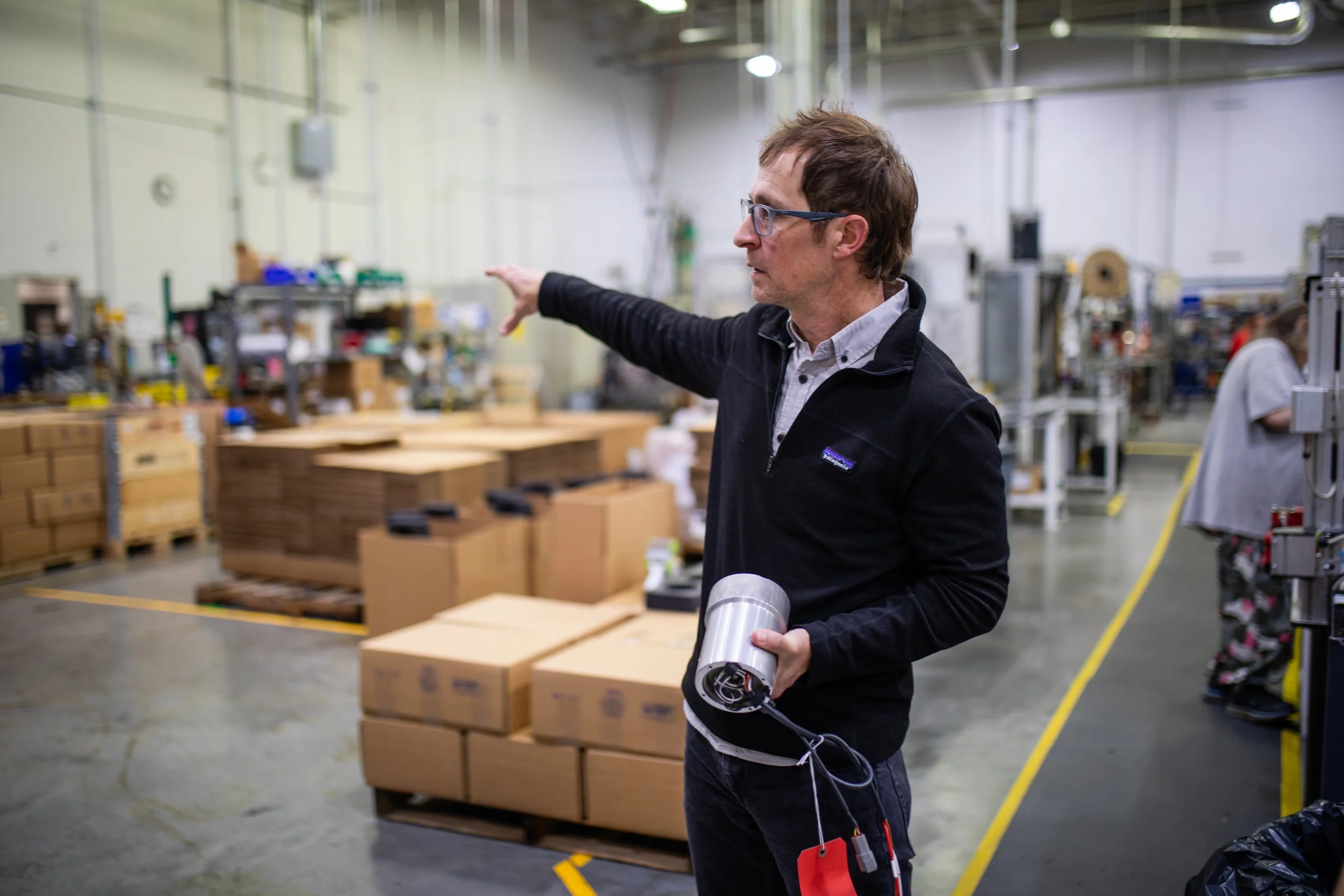 A man with glasses pointing with his right hand while holding a disassembled electrical motor in his left hand inside a warehouse or manufacturing facility.
