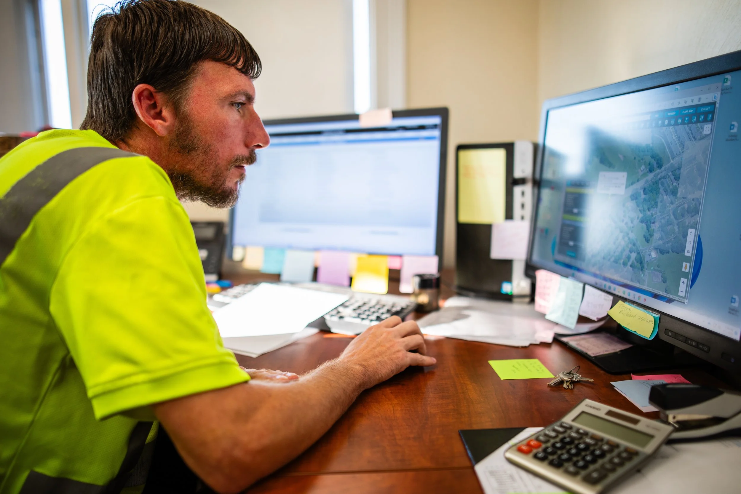 Man working at a desk with two computer monitors, one displaying a map, in an office with sticky notes and office supplies.