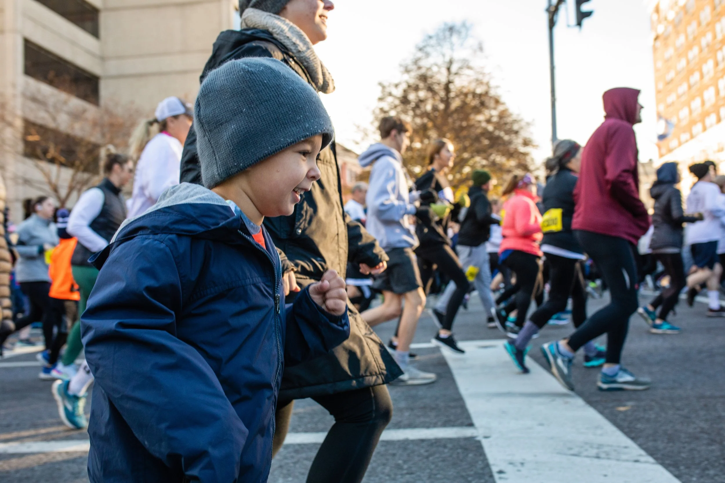 Young boy and adults running in a street race during the daytime, with a diverse group of people running in the background.