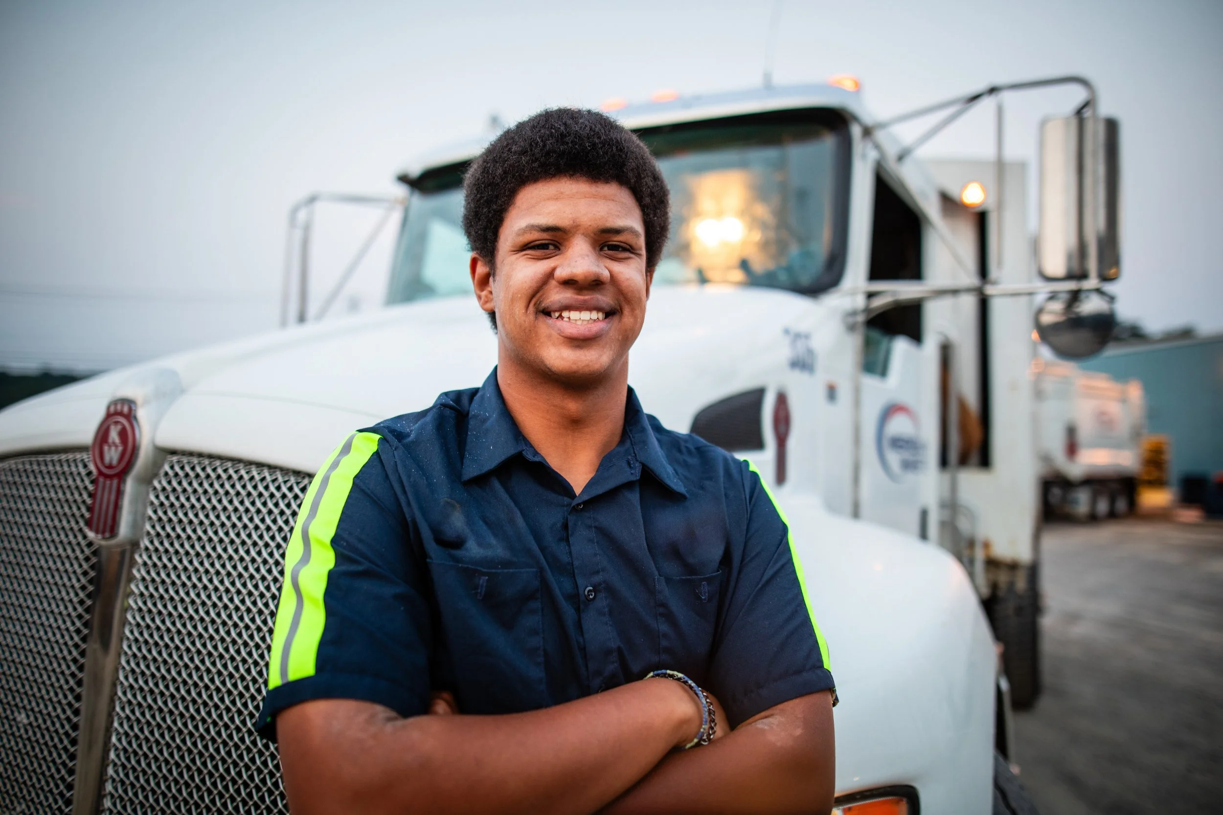 A young man with arms crossed smiling in front of a white semi-truck on an outdoor lot during sunset.