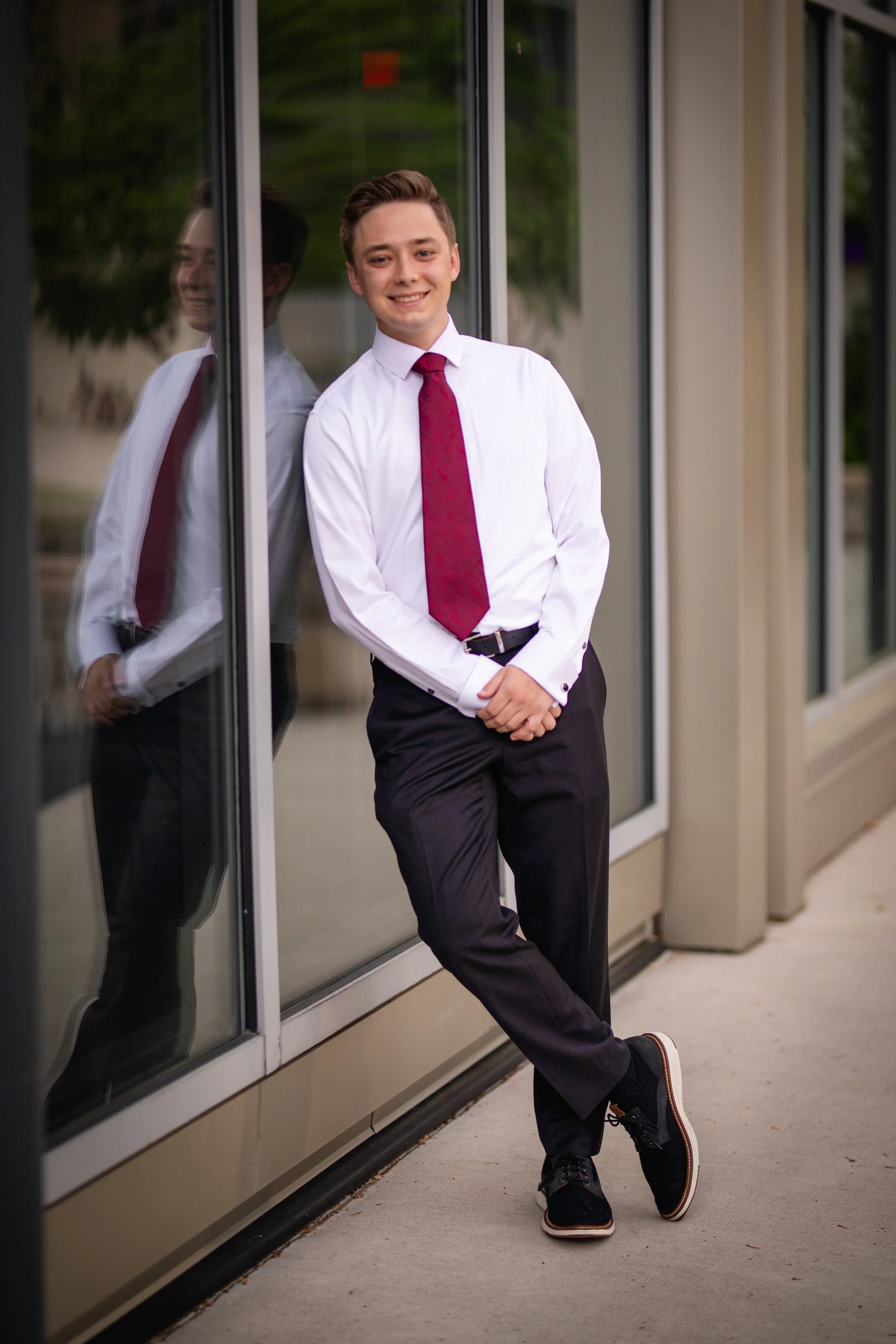 Young man in white shirt and red tie leaning against glass wall outside building, with reflection visible in the glass.
