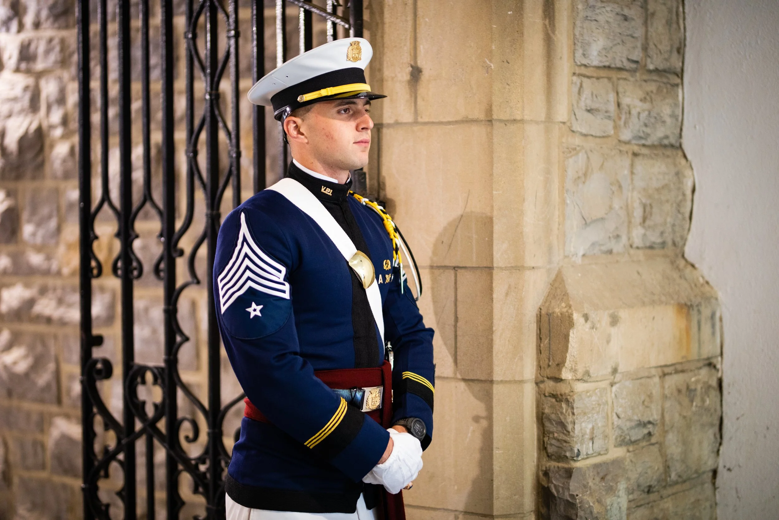 A young man in a ceremonial military uniform standing guard inside a building with stone walls and a black wrought iron gate.