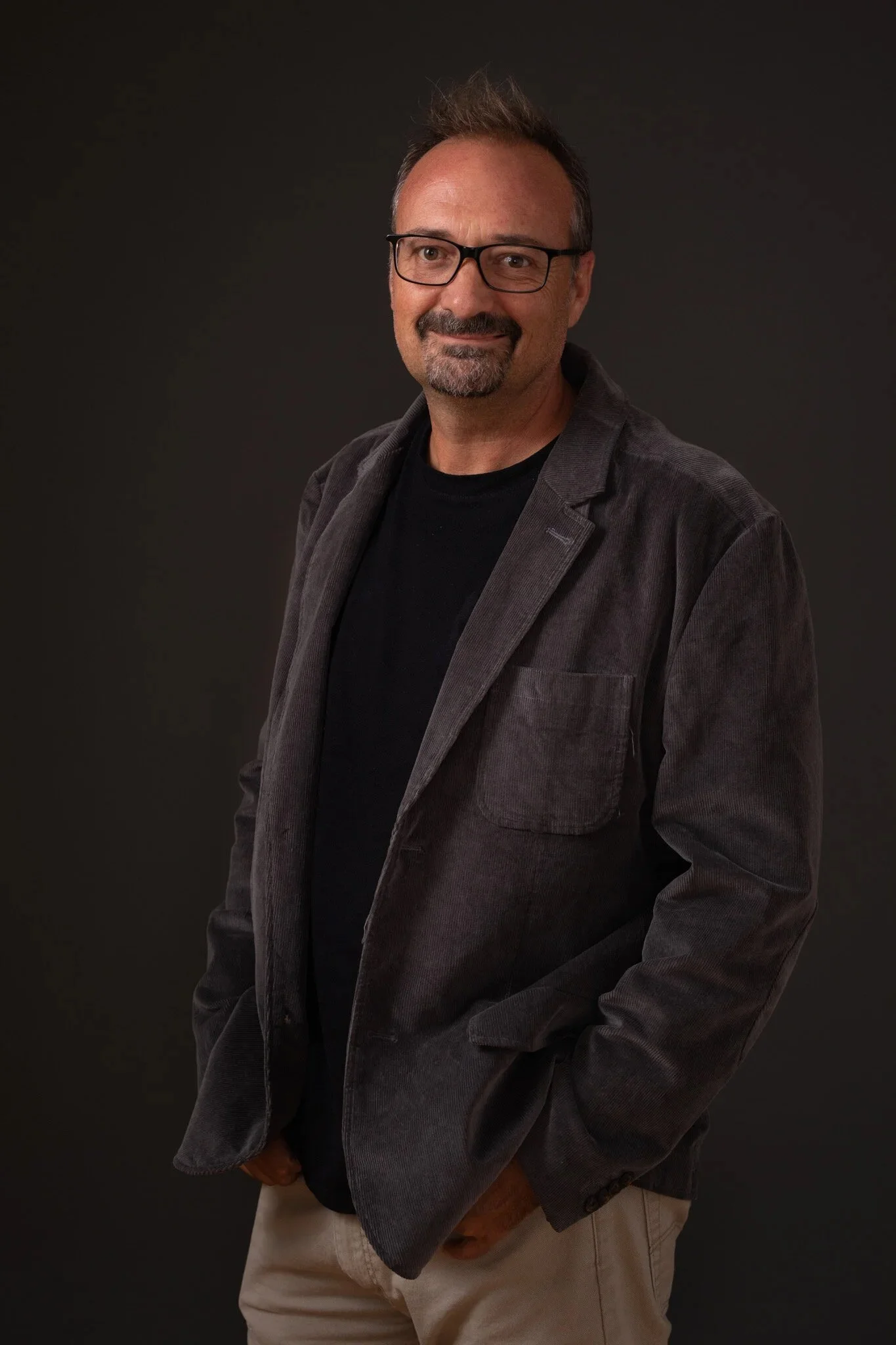 A middle-aged man with glasses, dark hair, and a beard, smiling and wearing a dark jacket over a black shirt and beige pants, standing against a dark backdrop.