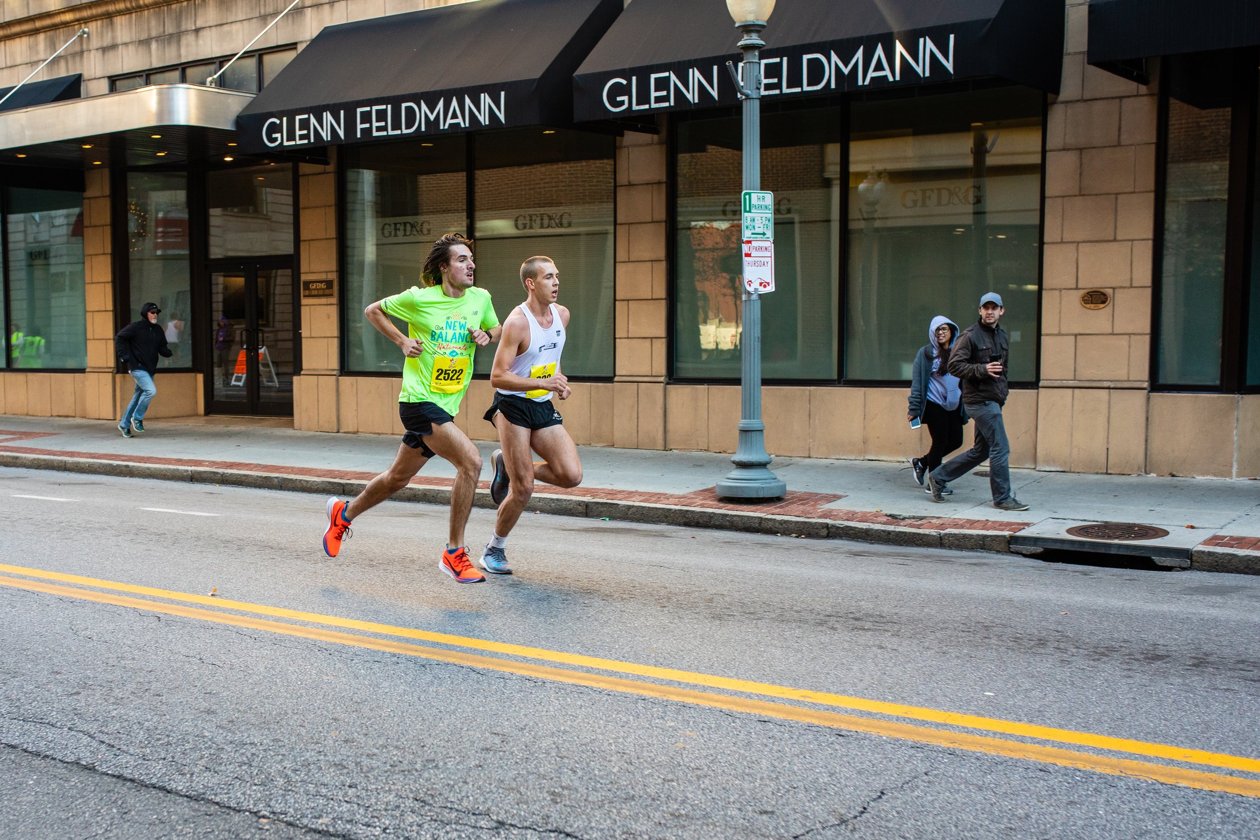 Two male runners participating in a race running on a city street, with spectators walking on the sidewalk and a storefront with black awnings and large windows in the background.