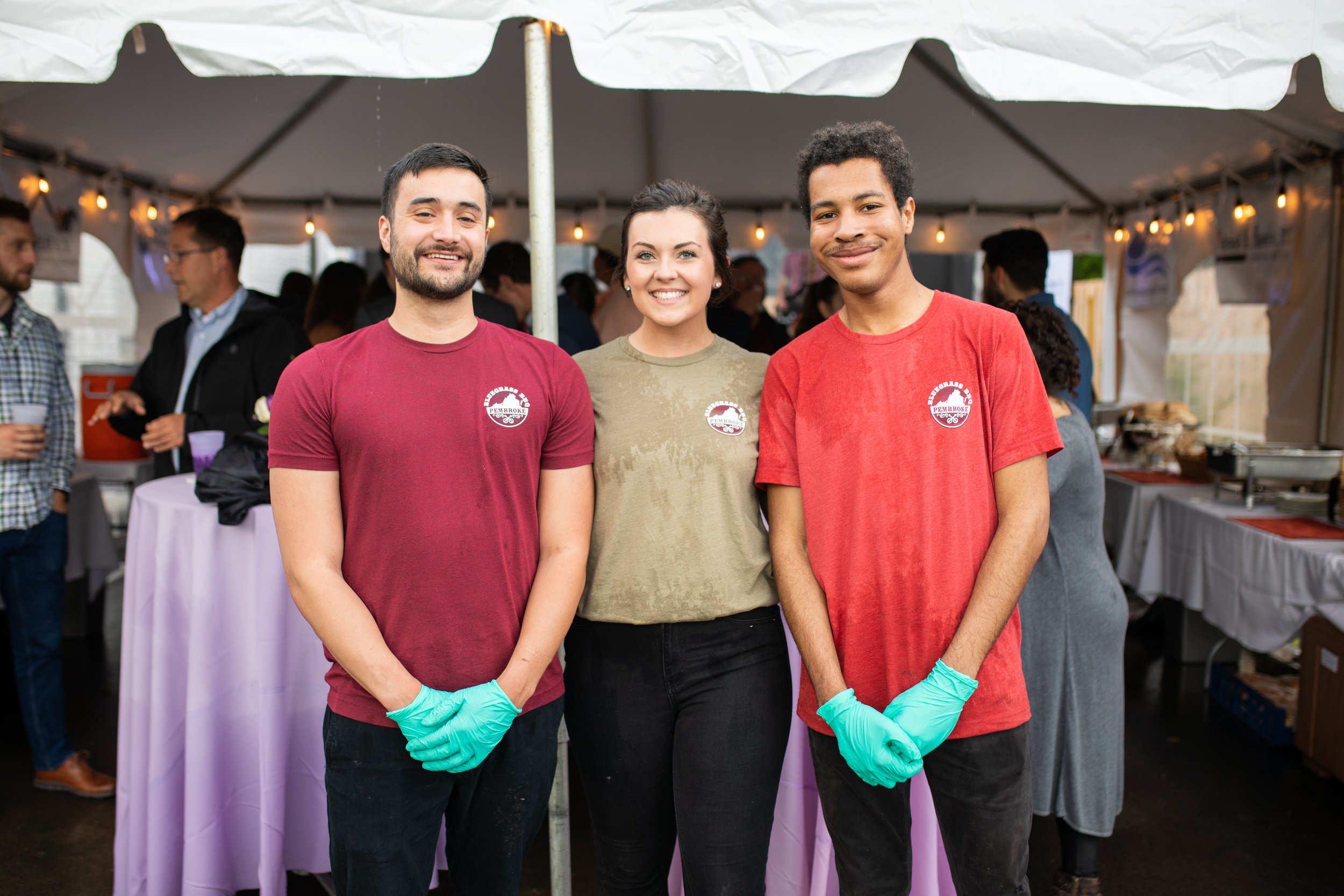 Three volunteers wearing gloves standing in front of a tent at a community event, smiling for the camera.