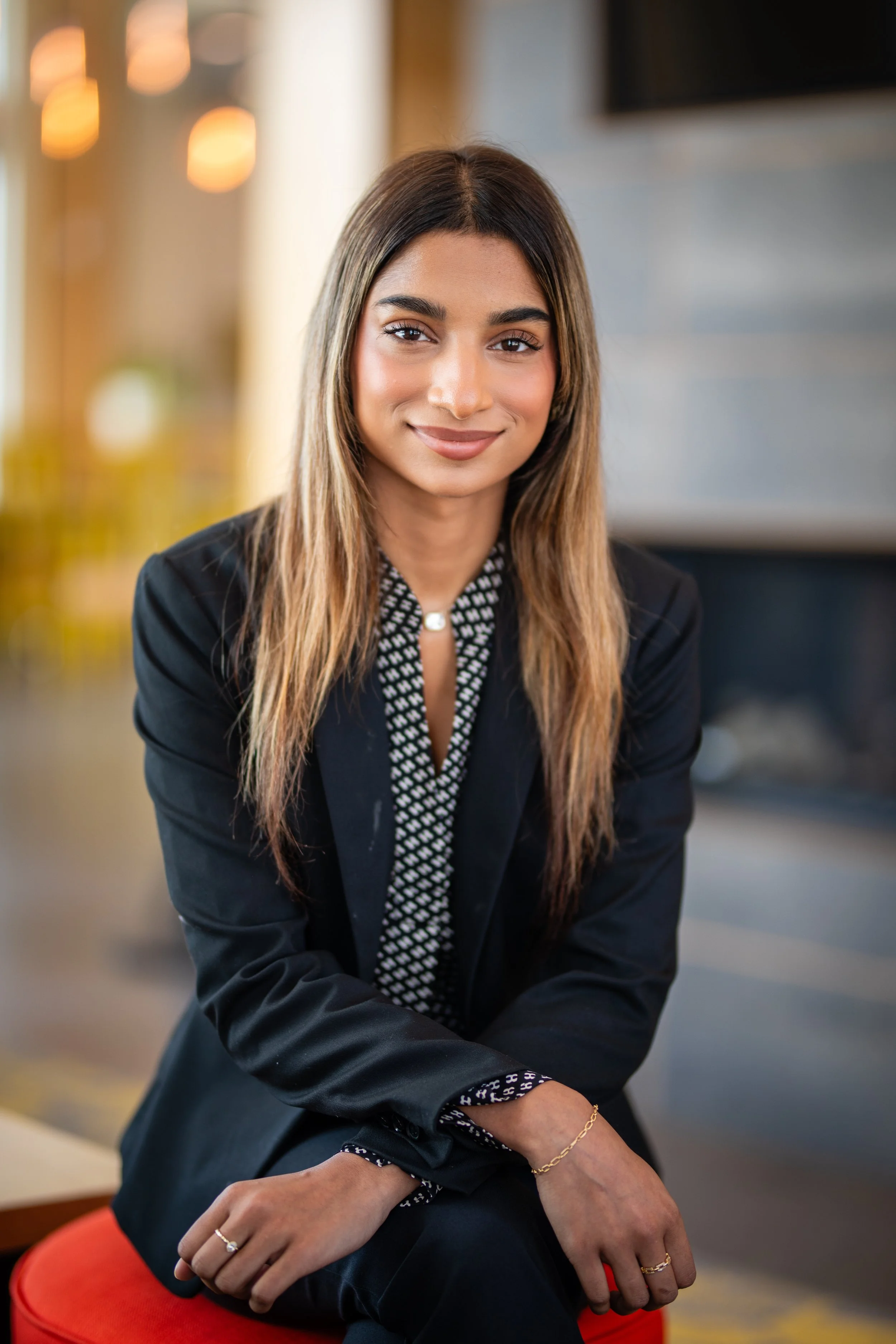 A young woman with long wavy hair, wearing a black blazer over a patterned black and white blouse, sitting and smiling in a warmly lit indoor setting.
