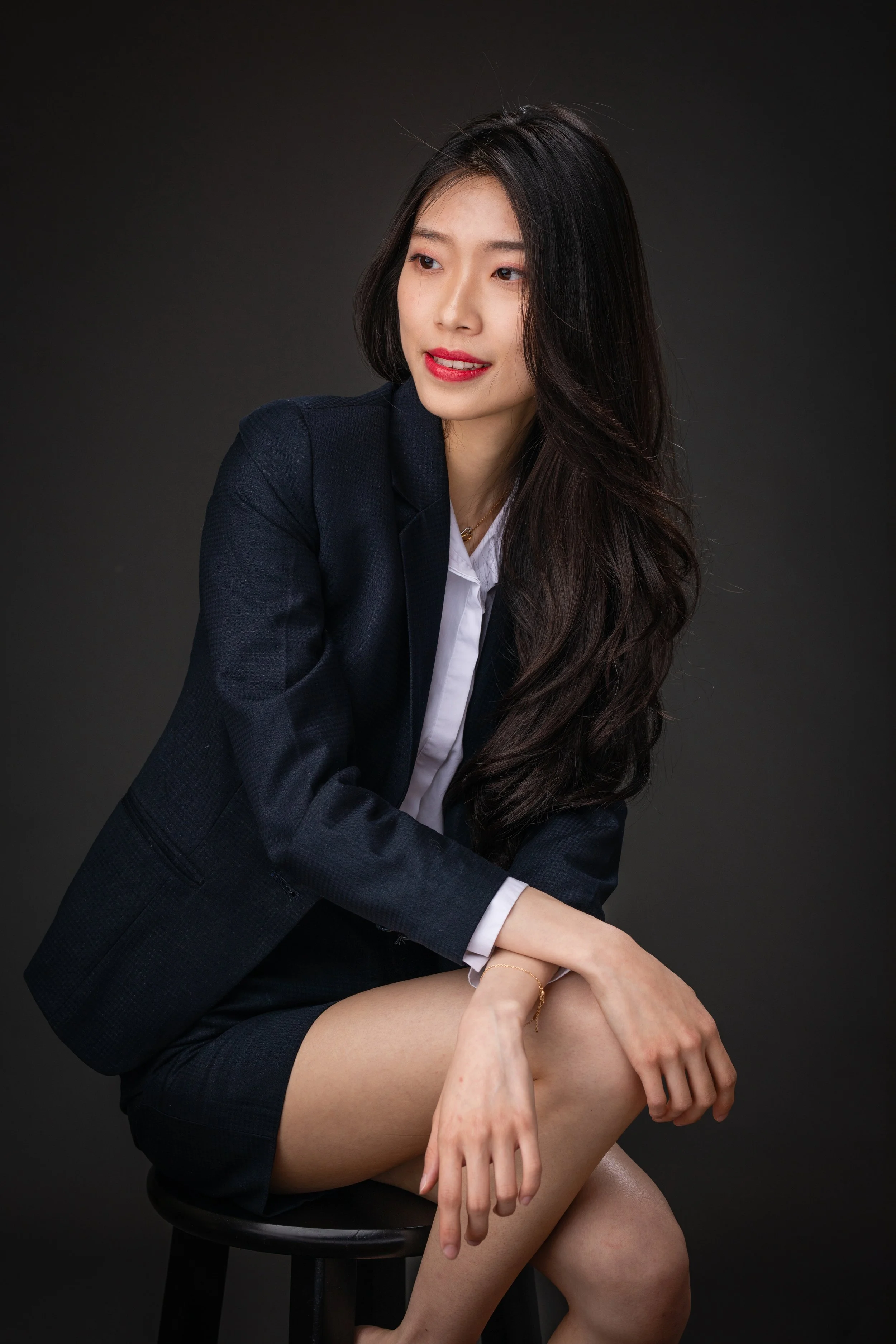 A woman sitting on a stool, wearing a dark blazer and white shirt, with long dark hair and red lipstick, against a dark background.