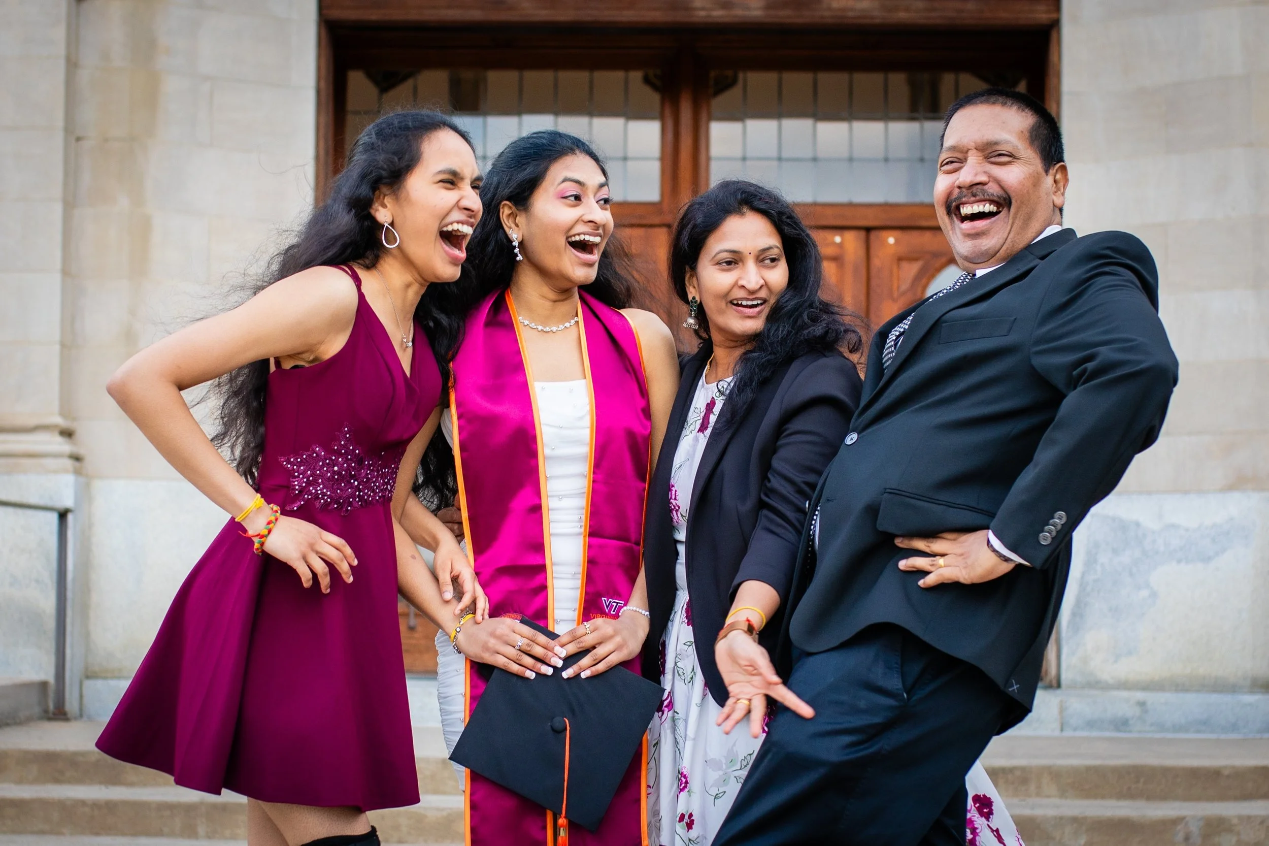 Four people, dressed formally, standing on stairs outside a building, laughing and smiling at each other. One woman is in a magenta dress, another is in a graduation gown and cap, another is in a black blazer over a floral dress, and the man is in a 