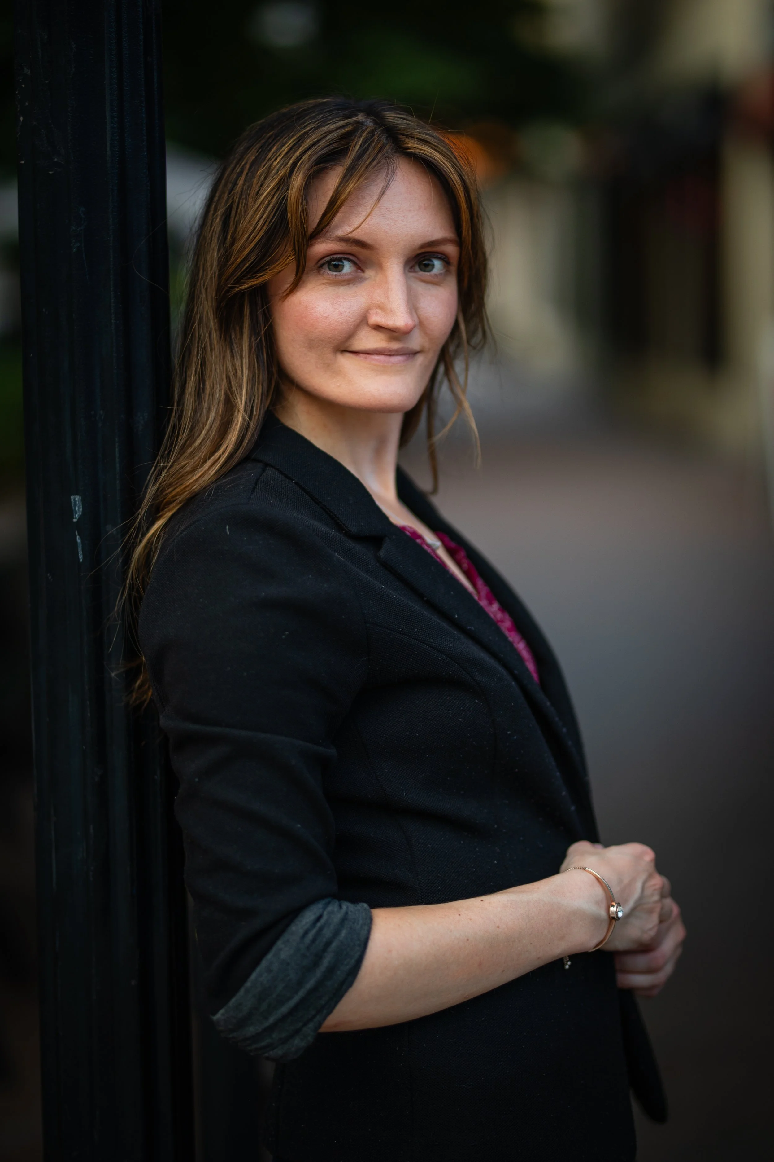 A woman with long, wavy brown hair, wearing a black blazer and pink top, leaning against a black pole outdoors during the evening, smiling softly.