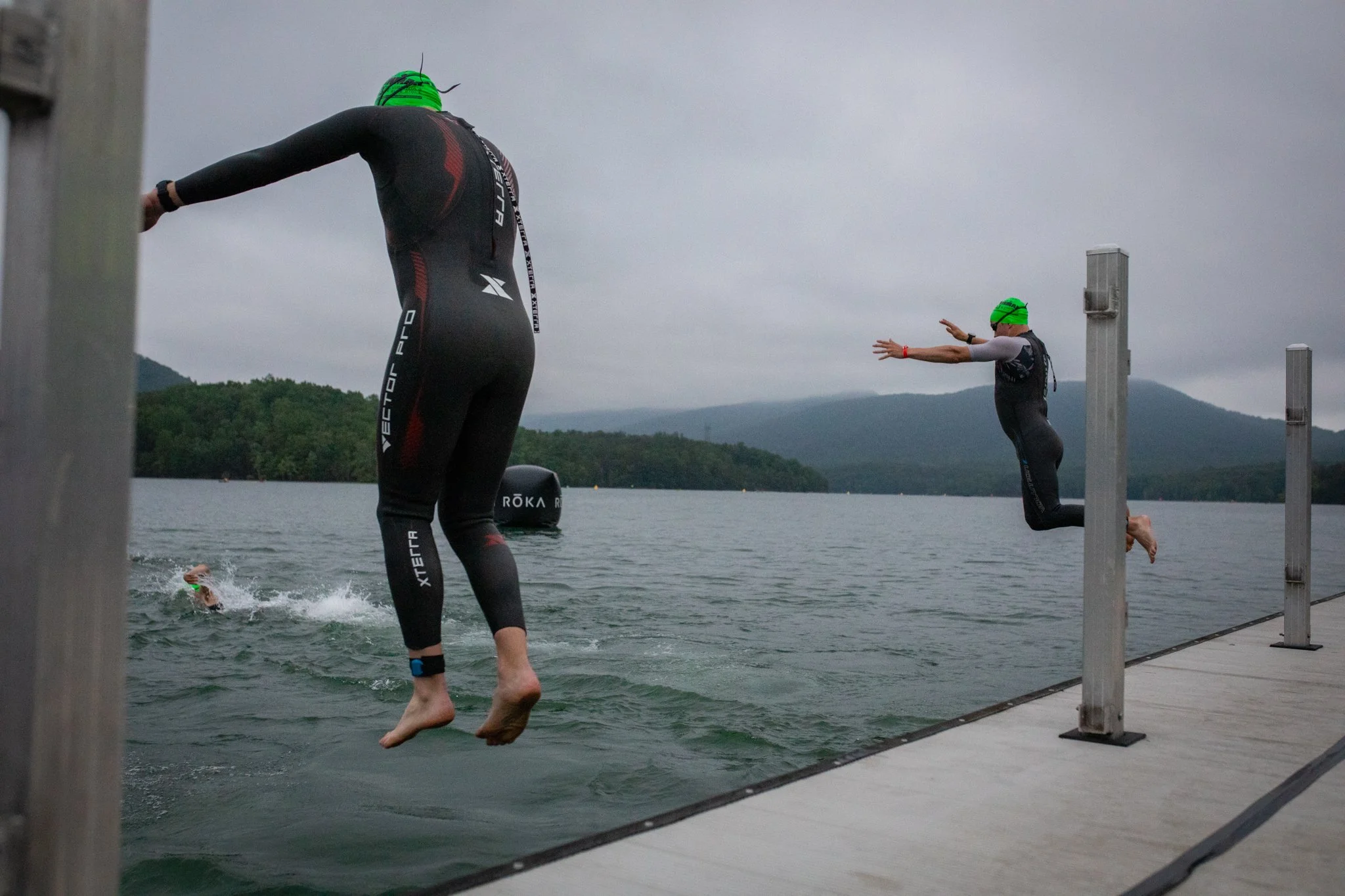 Two athletes in wetsuits and green swim caps jumping into a lake from a dock during a race or workout on an overcast day.