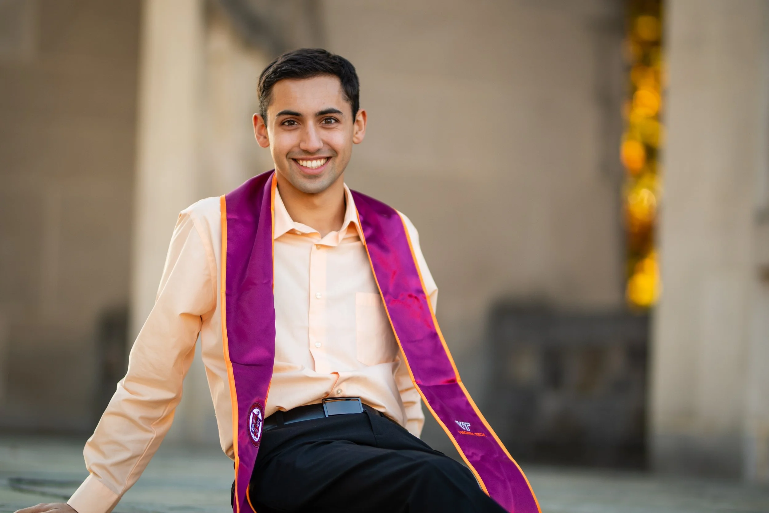 A young man in a light peach shirt and black trousers, smiling, wearing a purple graduation stole, standing outdoors.