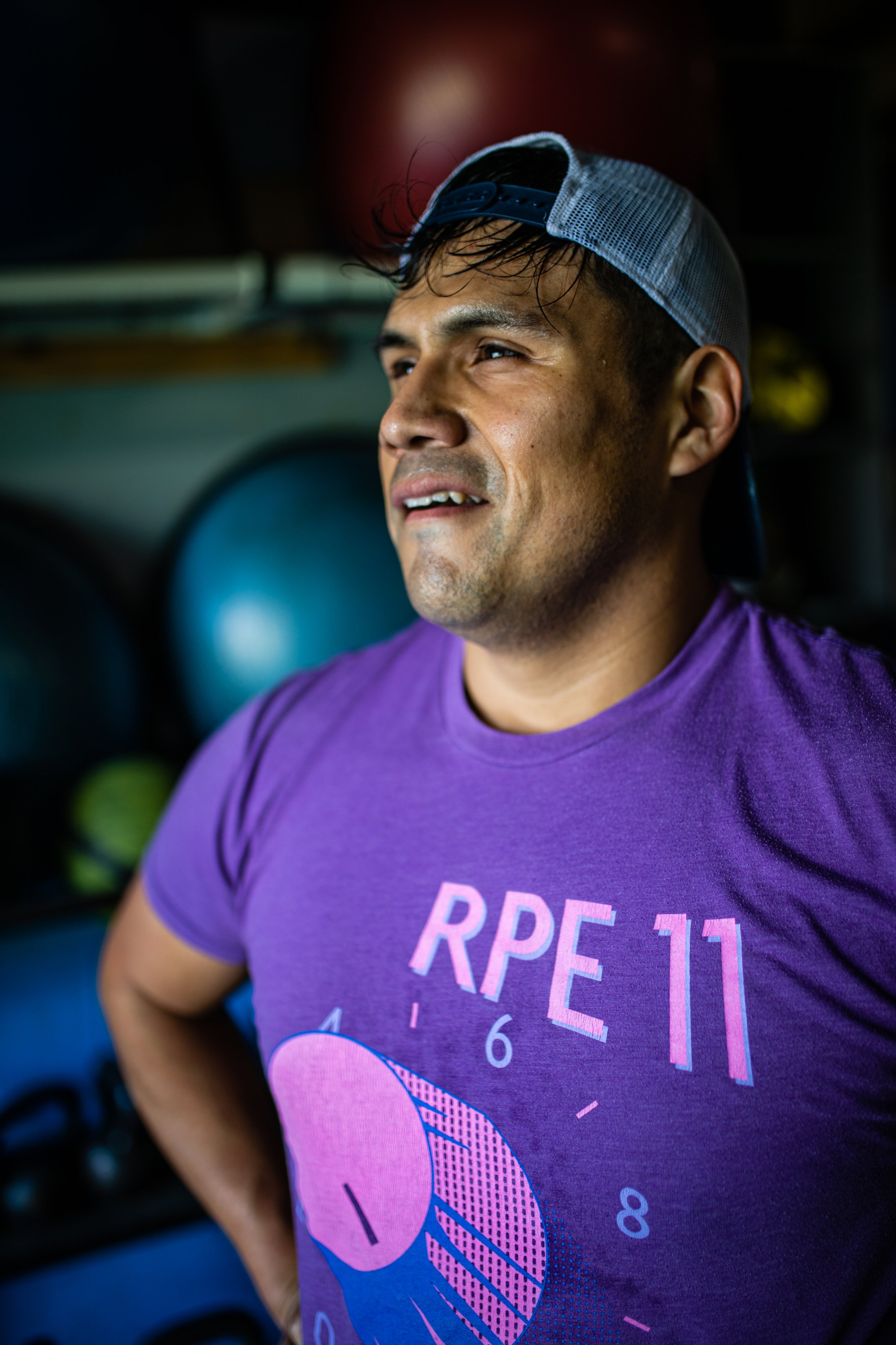A man wearing a purple RPE 11 t-shirt and a gray baseball cap backwards, standing in a gym with workout equipment in the background.