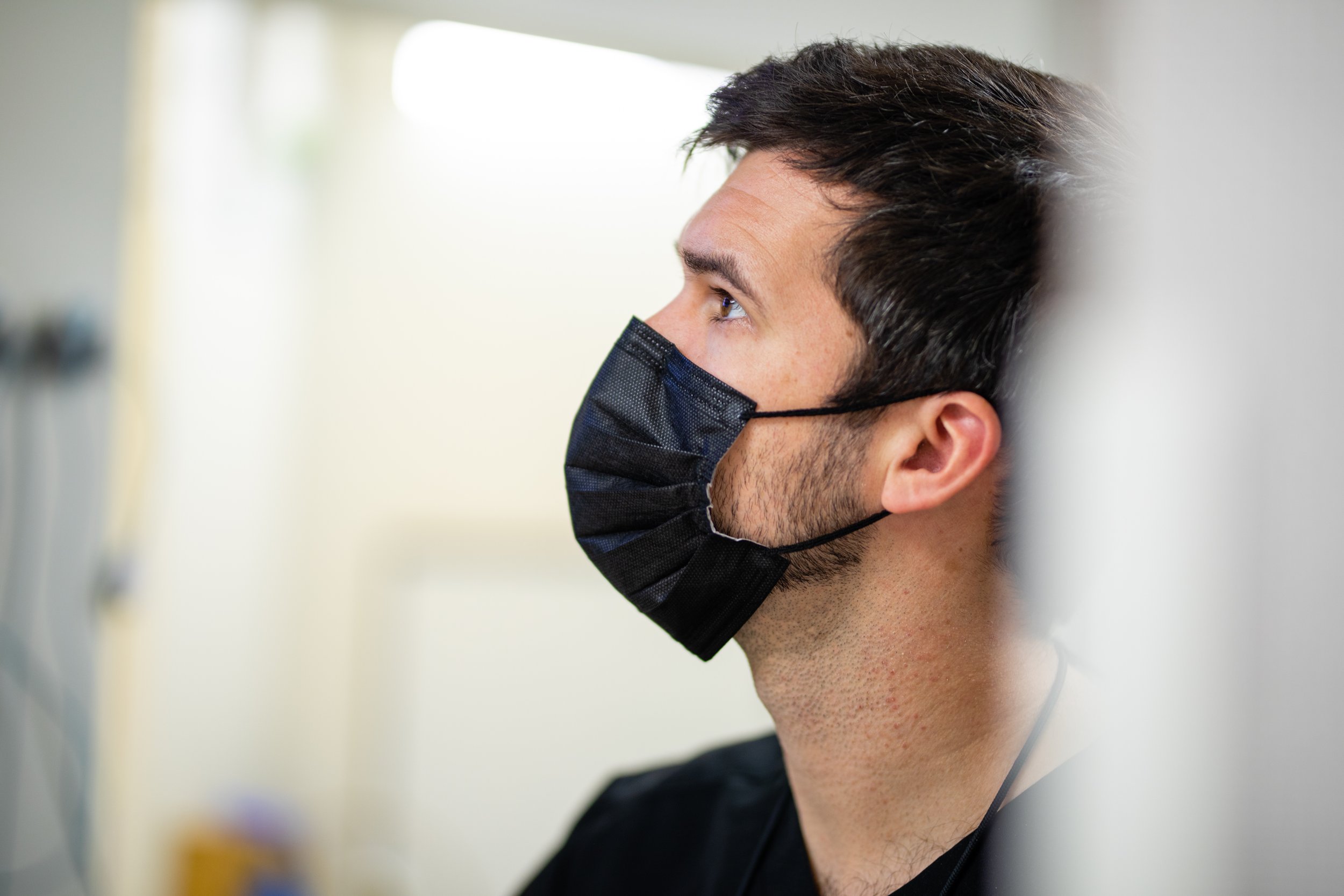 Side view of a man with dark hair and a beard wearing a black face mask, sitting indoors against a plain background.