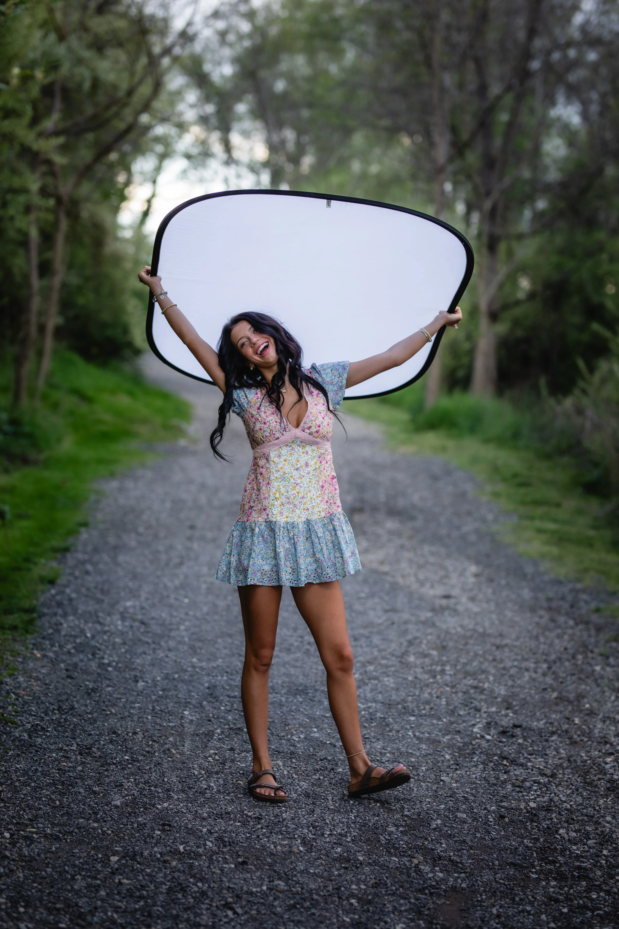 A young woman in a floral dress holding a large photography reflector outdoors on a gravel path surrounded by trees, smiling and posing cheerfully.