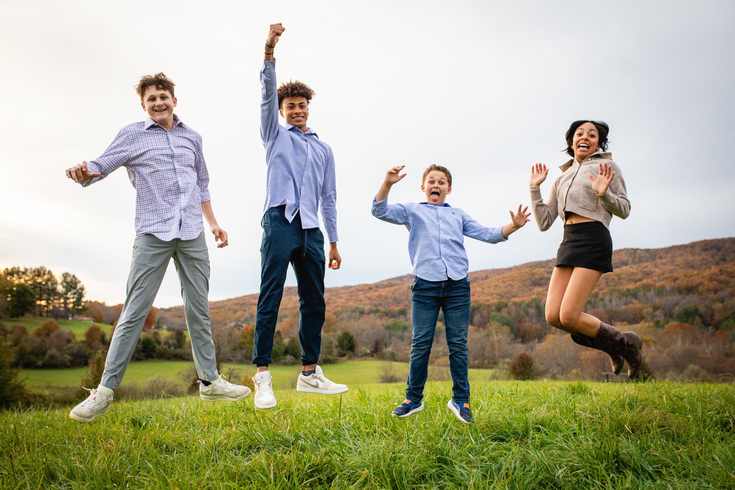 Four young people jumping in the air on a grassy field with rolling hills in the background, smiling and enjoying an outdoor activity.