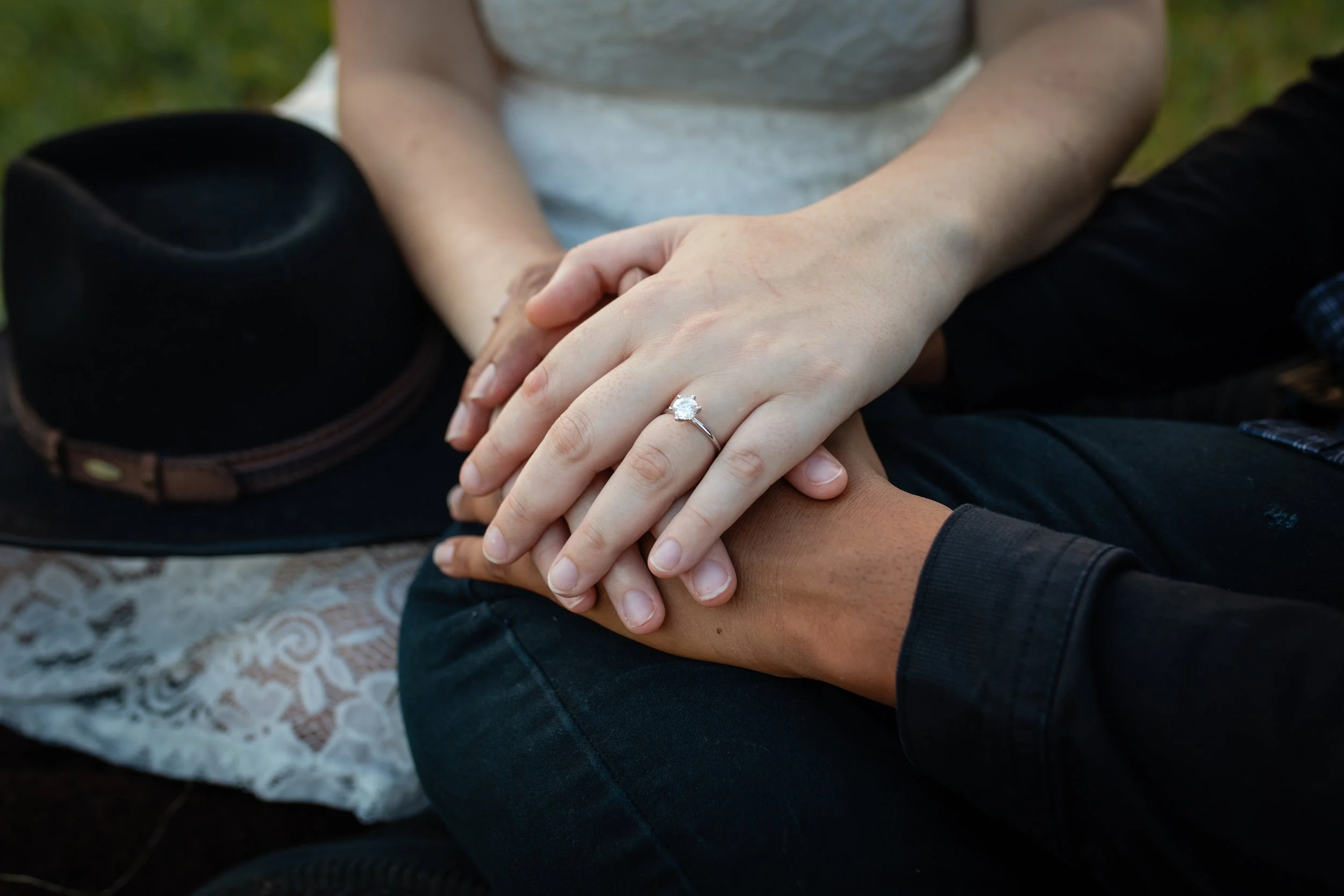 Close-up of two people holding hands, one wearing an engagement ring with a large diamond, outdoors with a hat on the ground beside them.