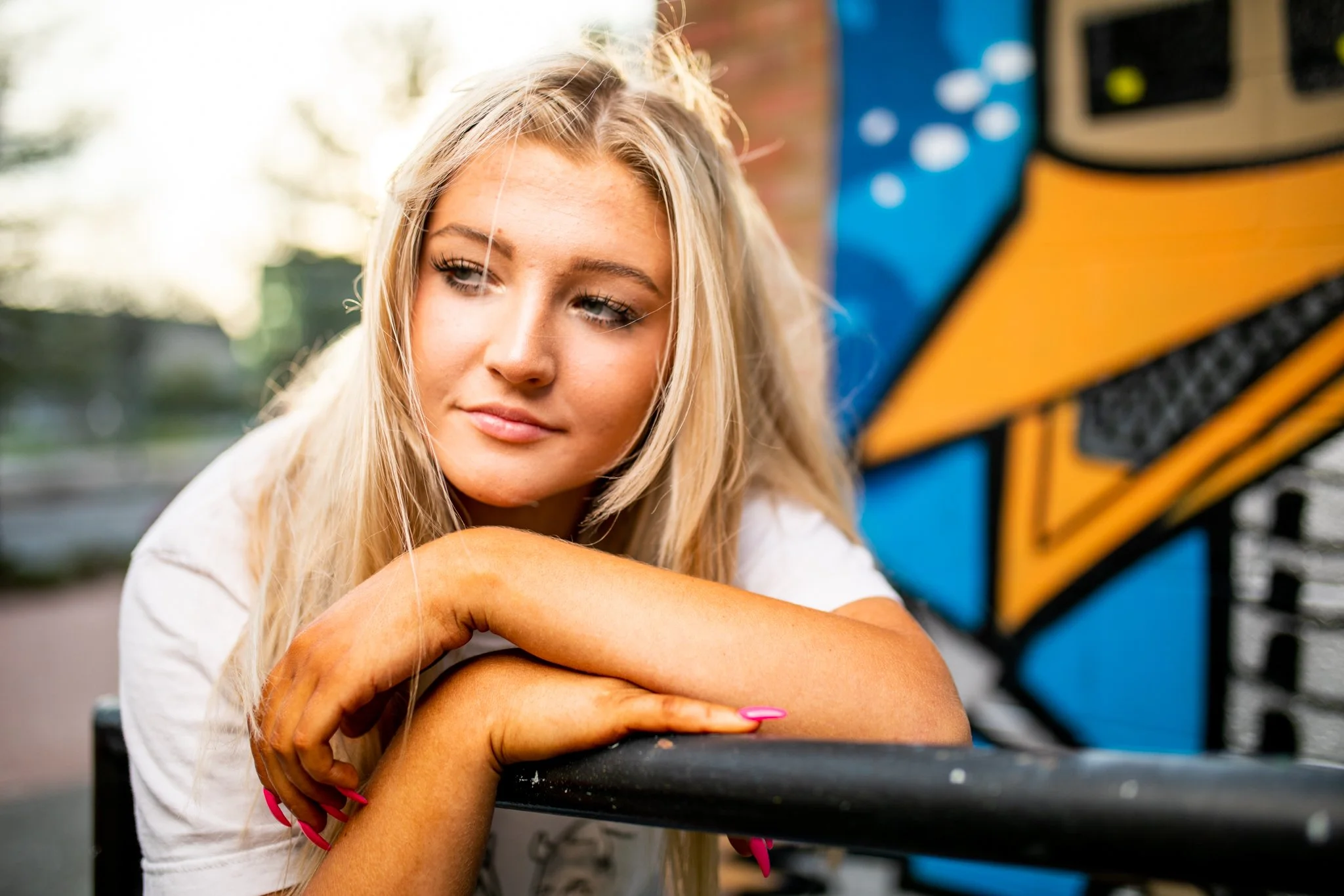 Young woman leaning on a black railing, looking to the side, with a colorful abstract mural in the background.