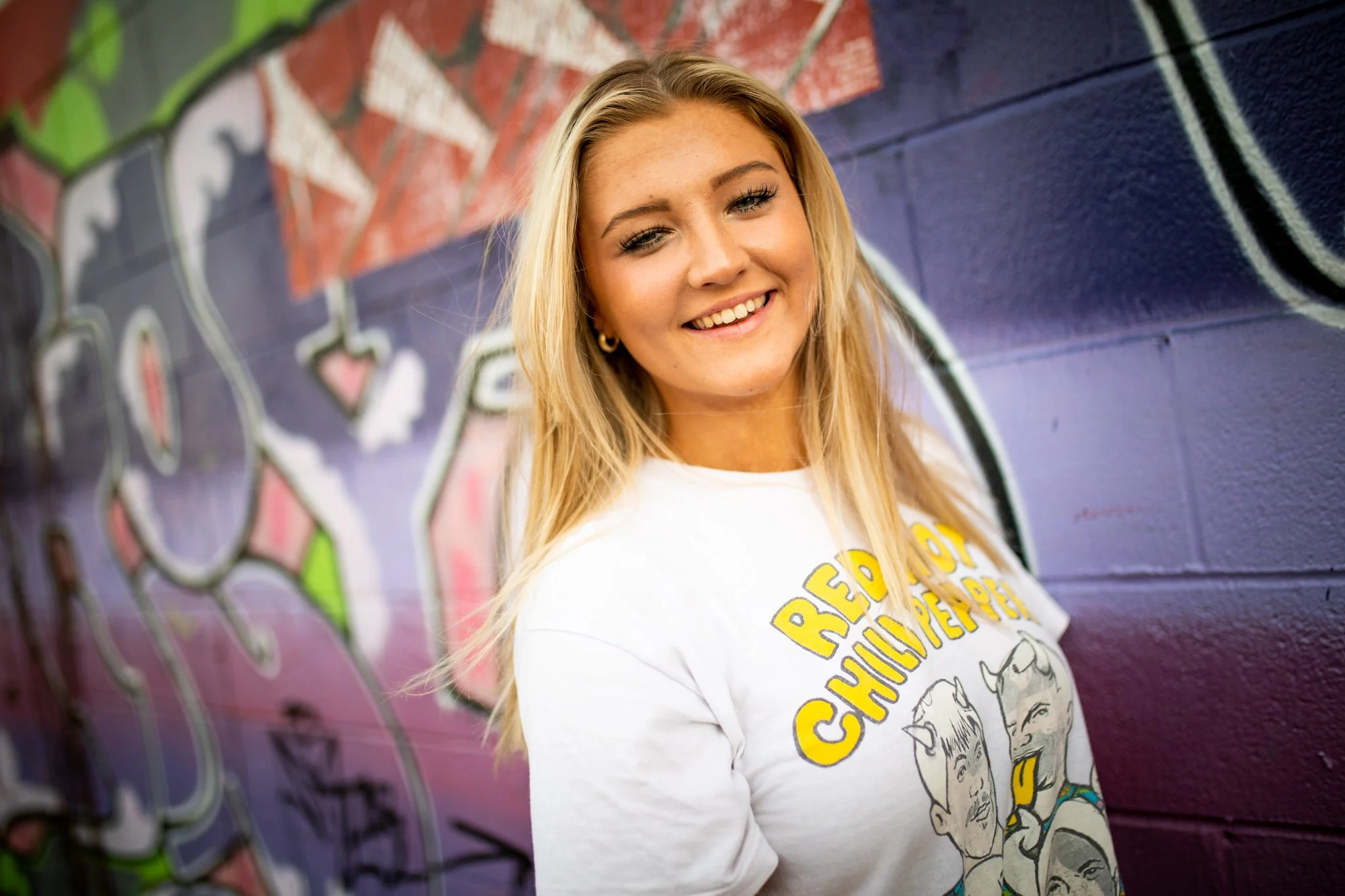 A young woman with blonde hair smiling in front of a colorful graffiti wall.