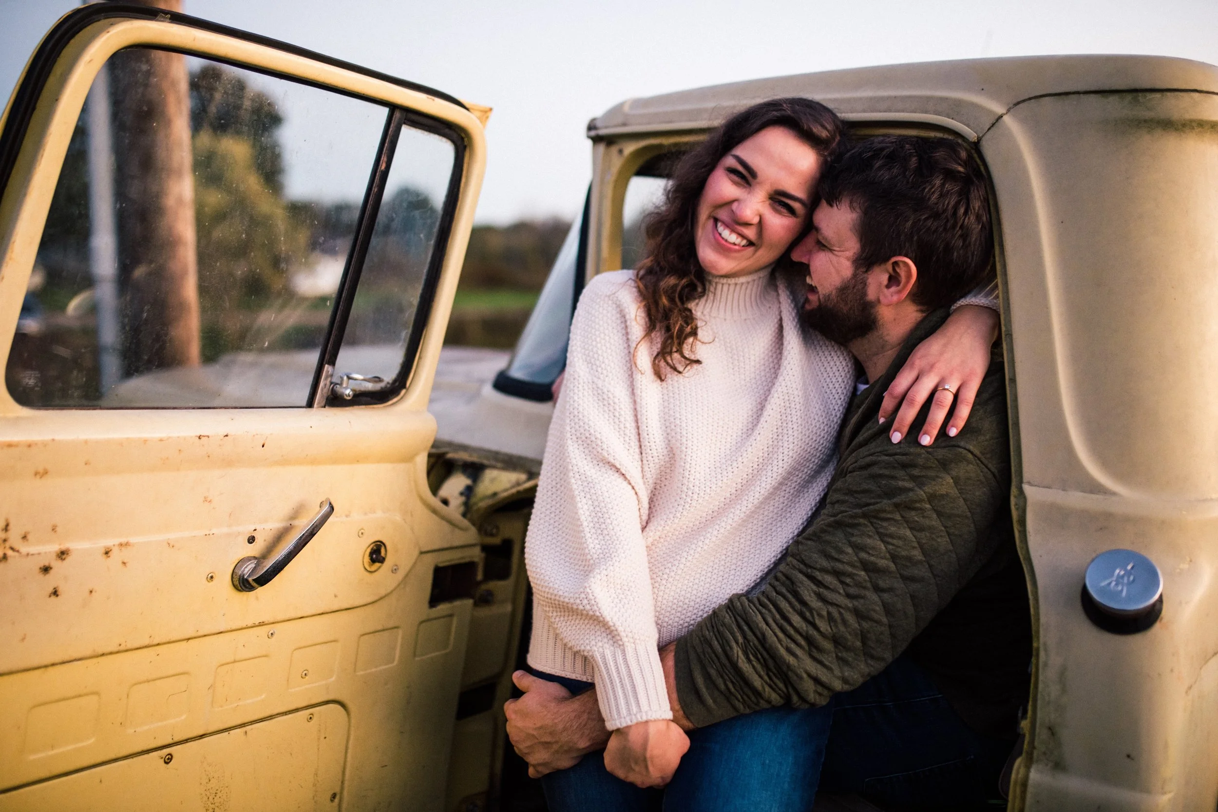 A happy couple embracing inside an old yellow vehicle, with the woman smiling and the man holding her by the waist, outdoors during sunset or late afternoon.