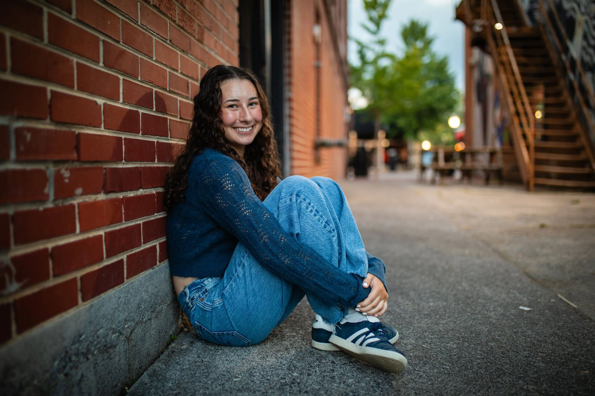 A young woman with curly hair sitting on the sidewalk next to a brick wall, smiling at the camera. She is wearing a blue sweater, blue jeans, and sneakers.