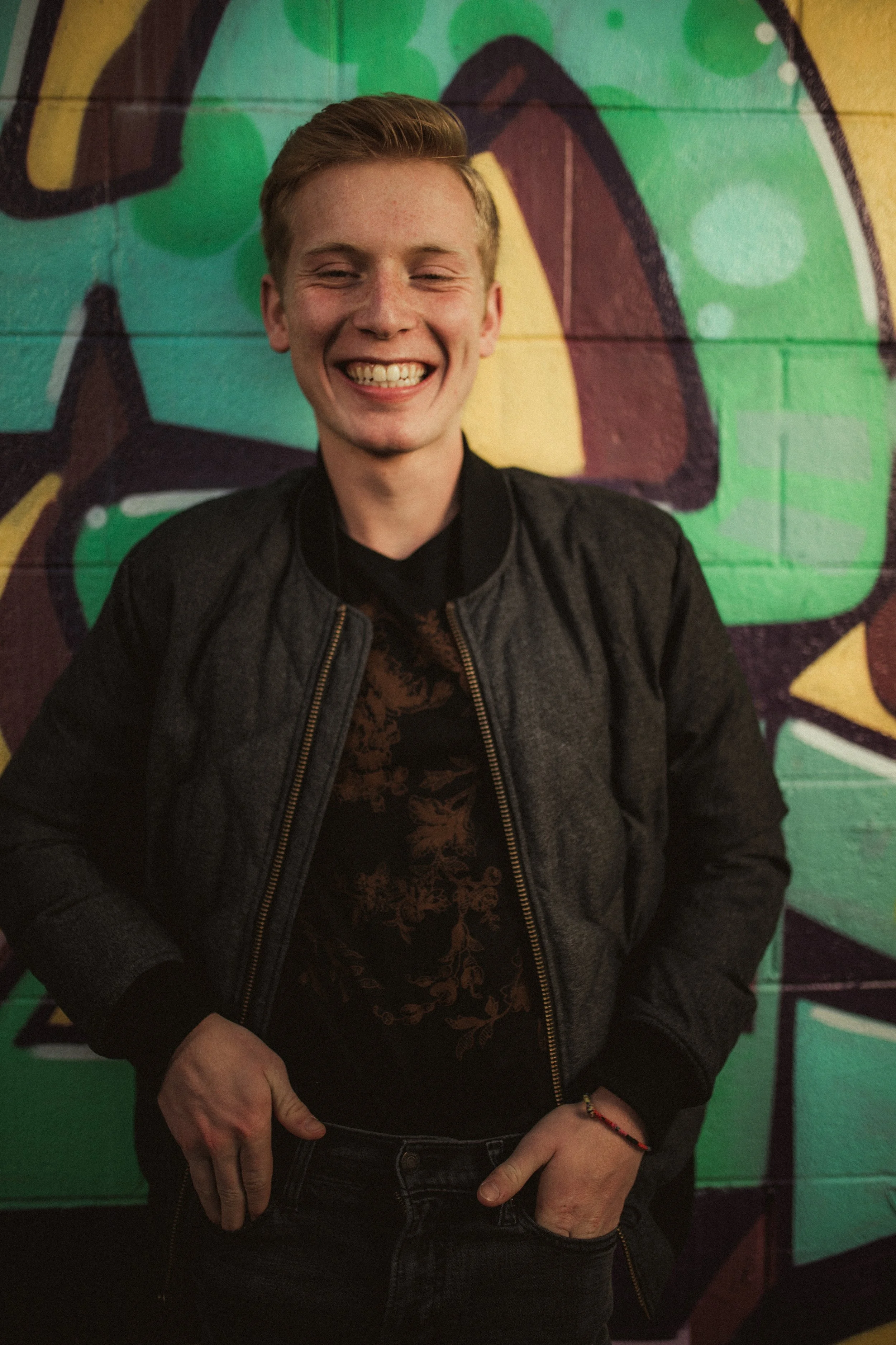 A young man with short blonde hair, smiling broadly, standing in front of a colorful graffiti wall.