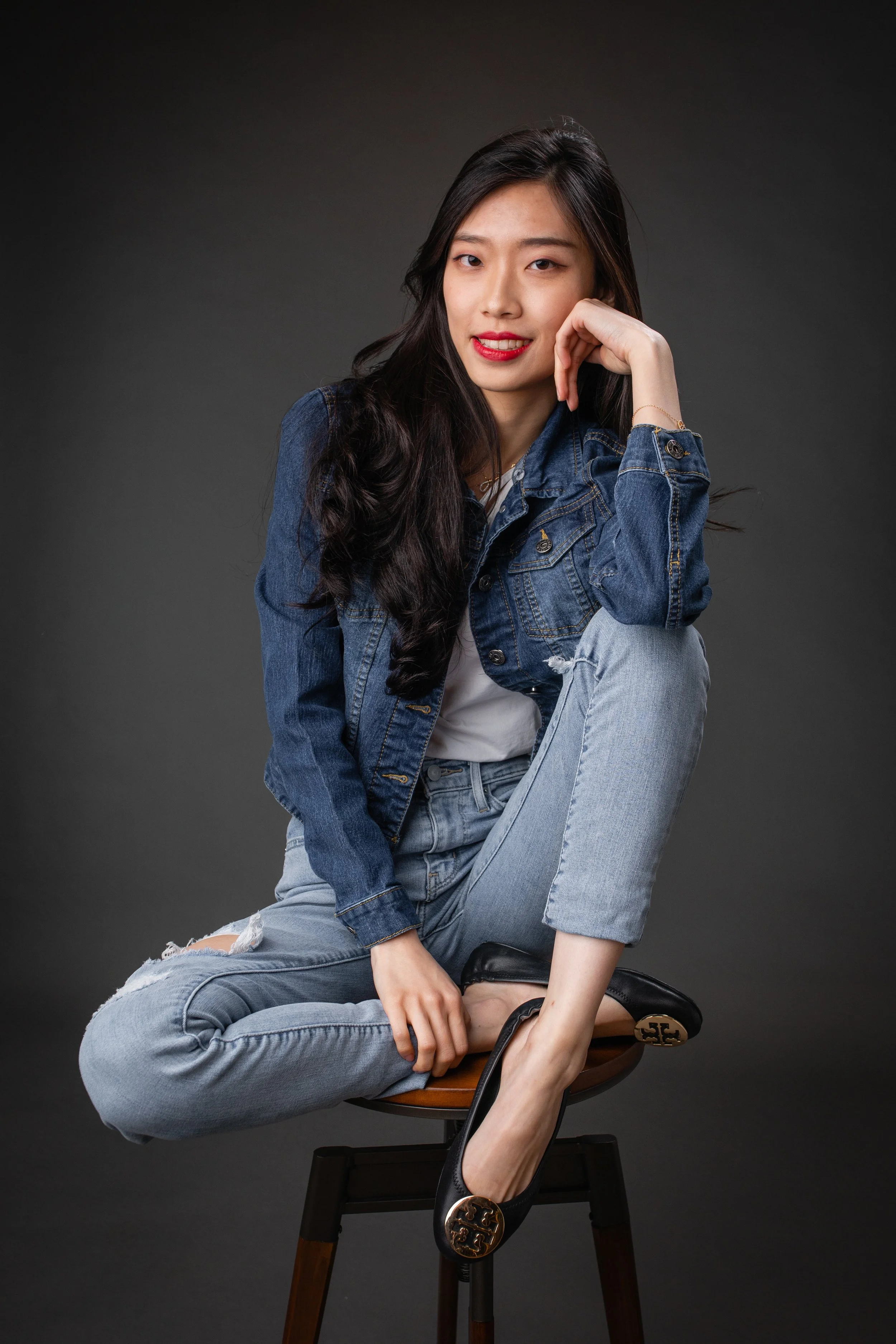 A young woman with long dark hair, wearing a denim jacket, white t-shirt, and ripped jeans, sitting on a stool against a dark background. She is looking at the camera, smiling with her hand resting near her face, and has one leg crossed over the othe