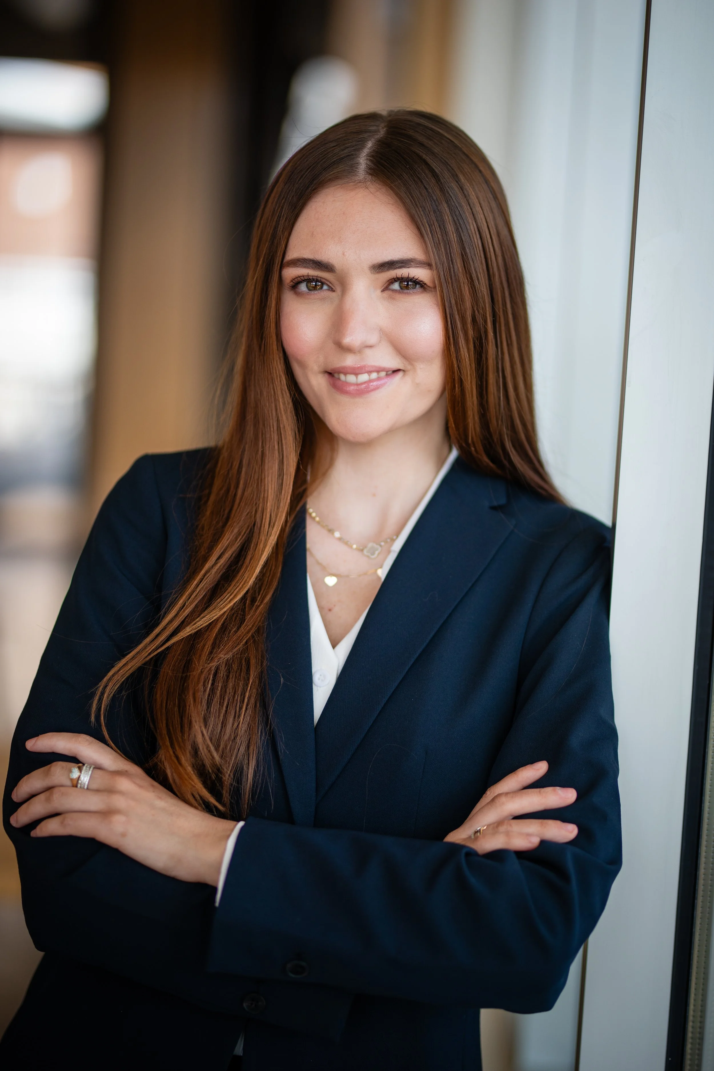 A young woman with long brown hair, wearing a navy blue blazer and white blouse, standing by a window with her arms crossed and smiling.
