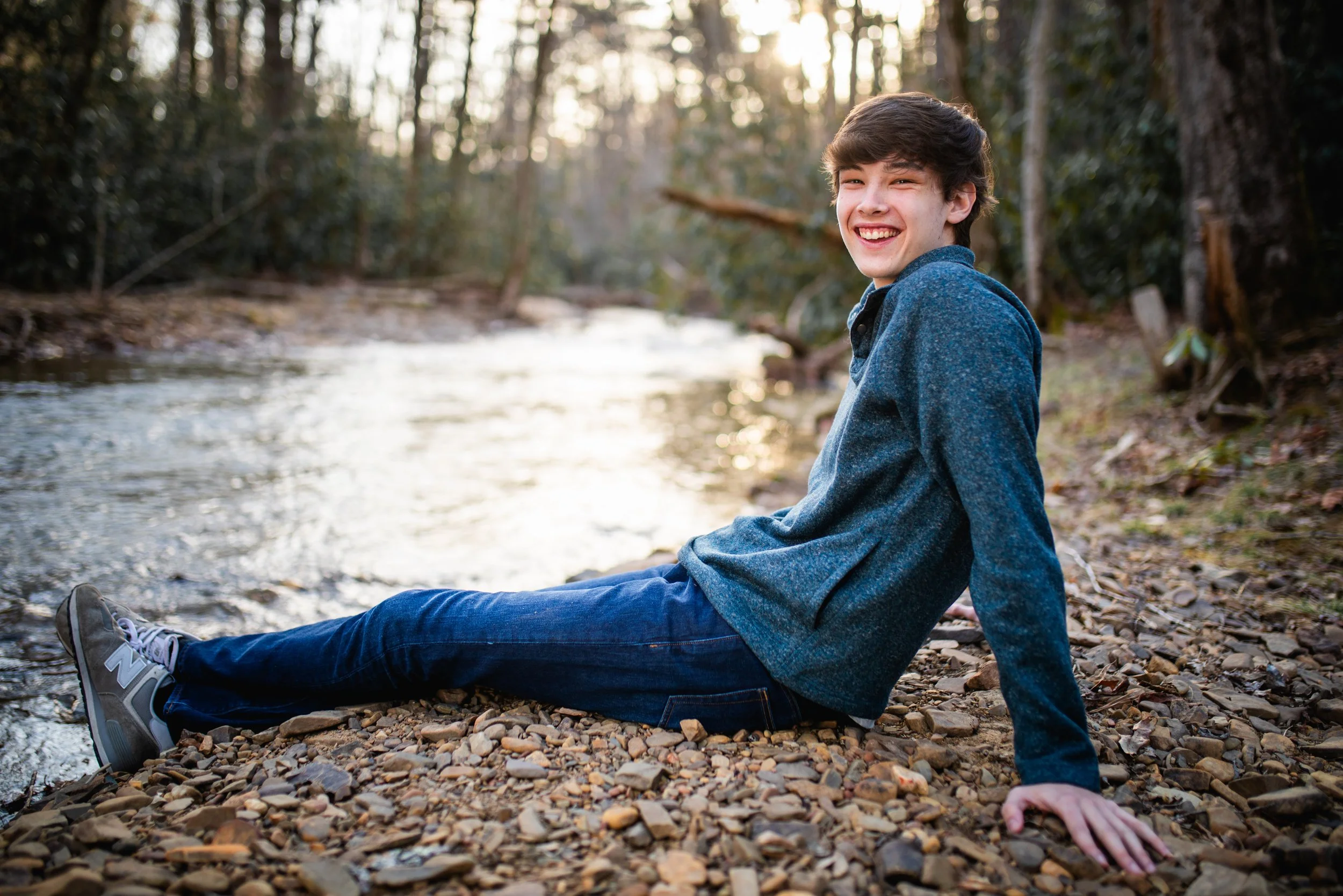 A young man sitting on a rocky riverbank, smiling, near a flowing river surrounded by trees during sunset.