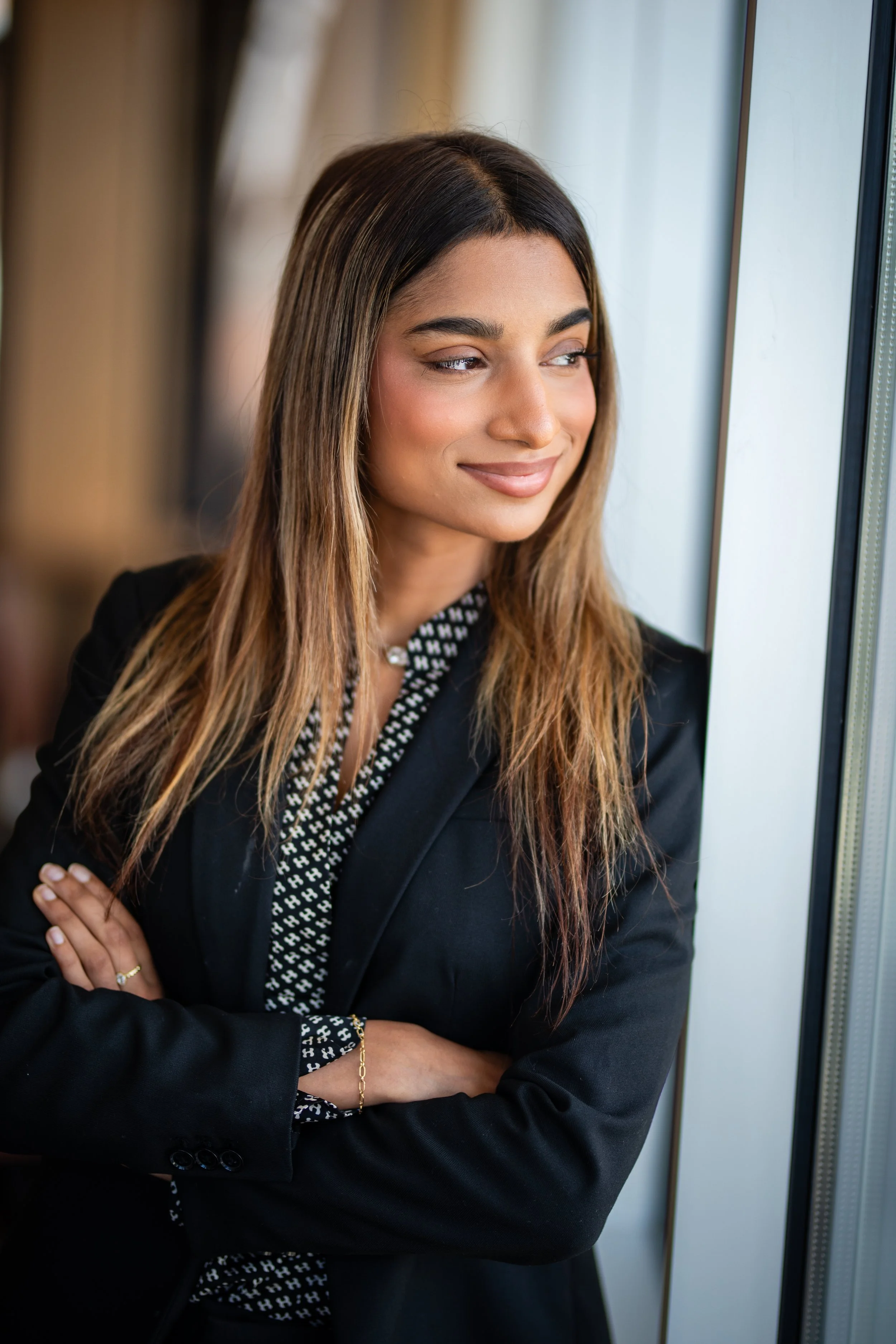 A woman with brown hair looking out a window, arms crossed, wearing a black blazer and patterned blouse.