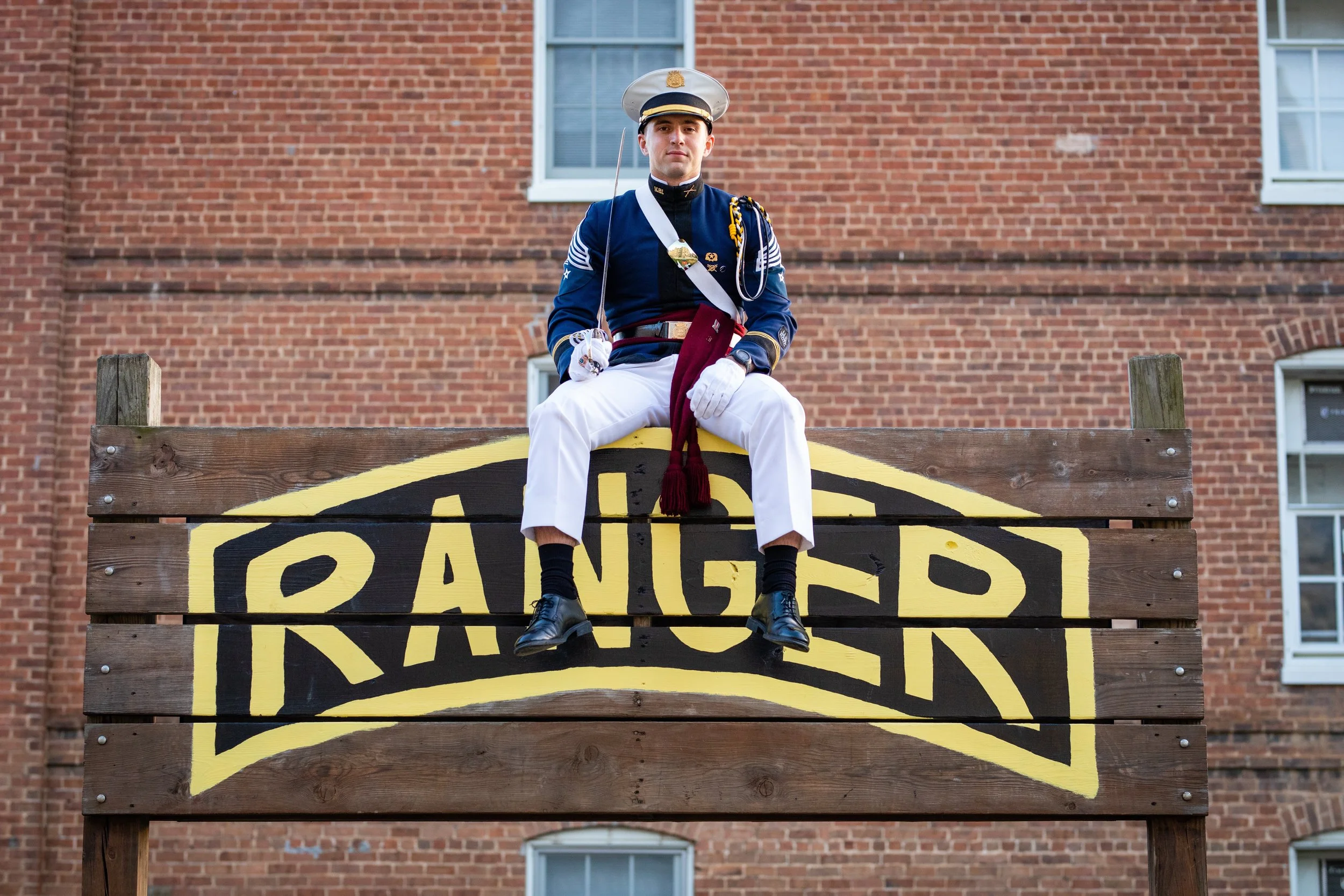 Person dressed in a military uniform sitting on a wooden sign painted with the word 'RANGER' in yellow and black, with a brick building in the background.