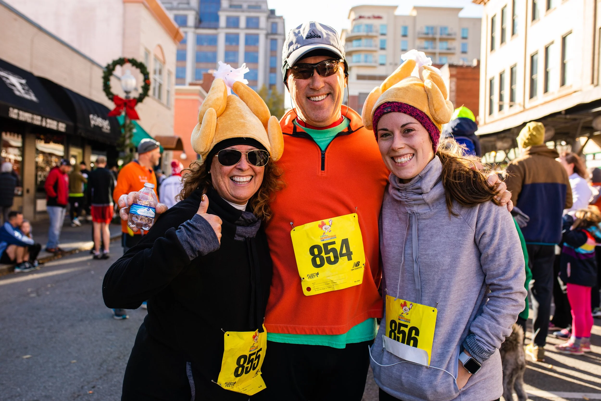 Three people wearing turkey hats and race bibs, smiling at an outdoor event with city buildings in the background.