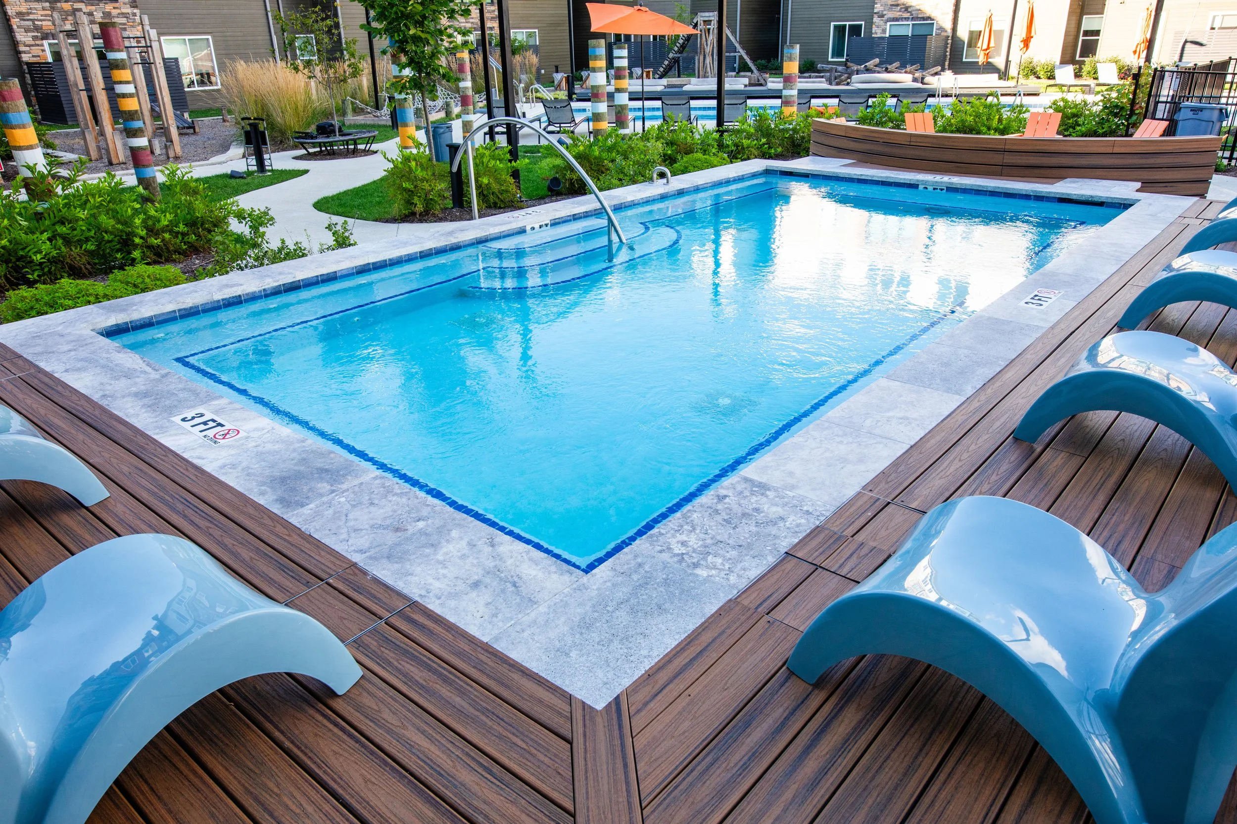A rectangular swimming pool surrounded by a wooden deck with lounge chairs, greenery, umbrellas, trees, and modern outdoor furniture in a residential complex.