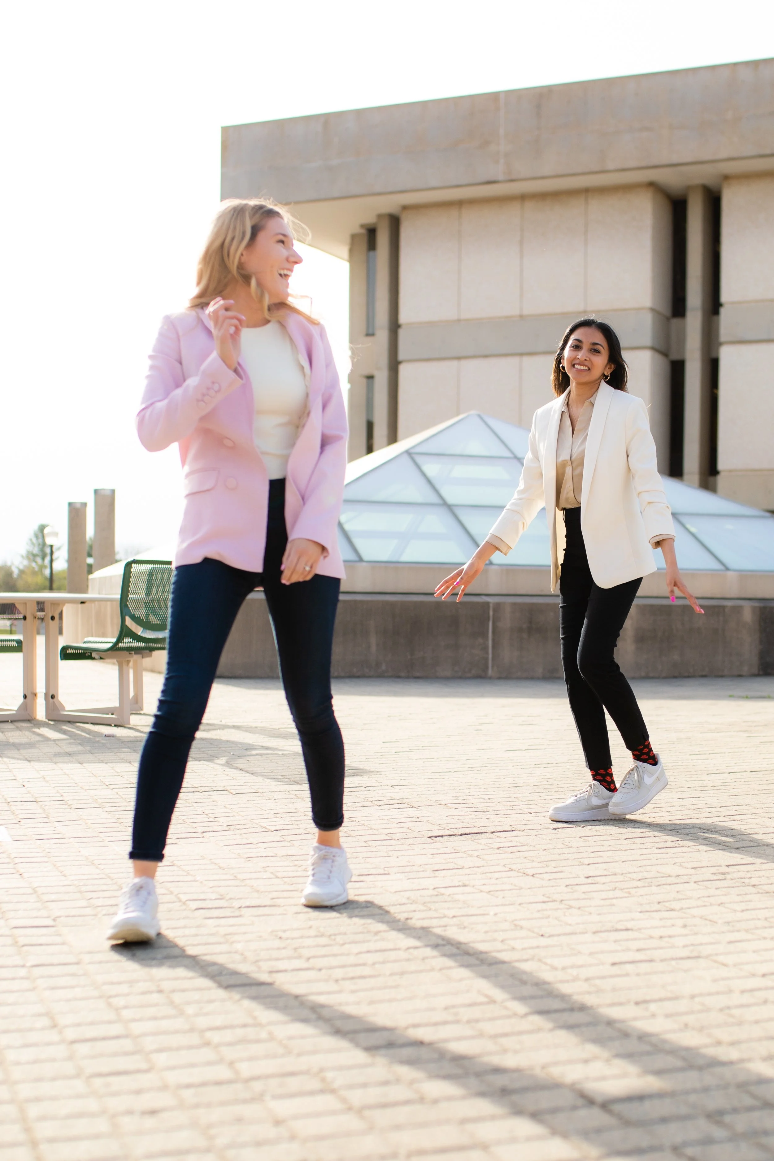 Two women stand outdoors on a paved area near a modern building, smiling and interacting playfully with each other.