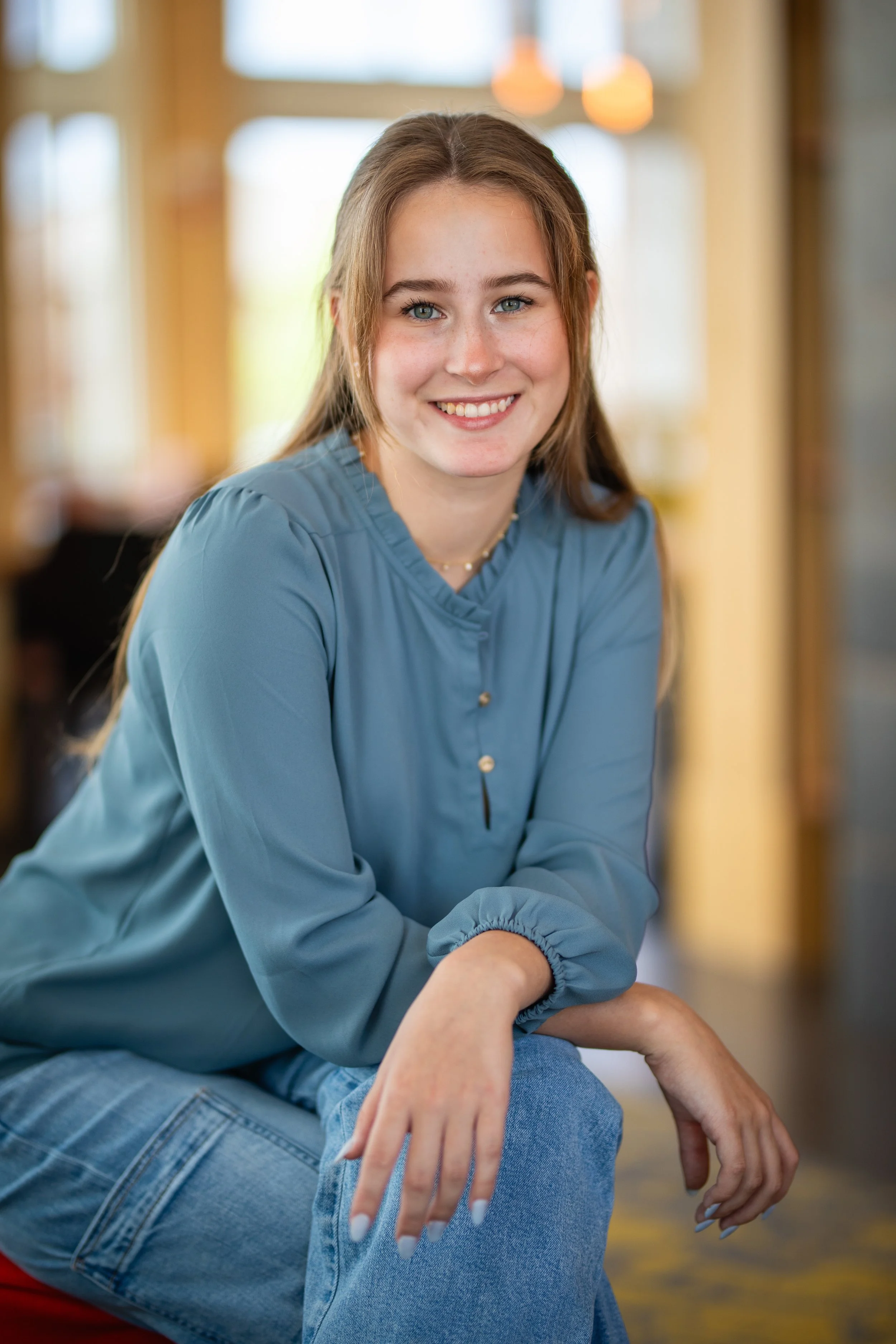 A young woman with long brown hair, blue eyes, and a bright smile, wearing a blue blouse and denim jeans, sitting indoors with a blurred background of warm lighting.